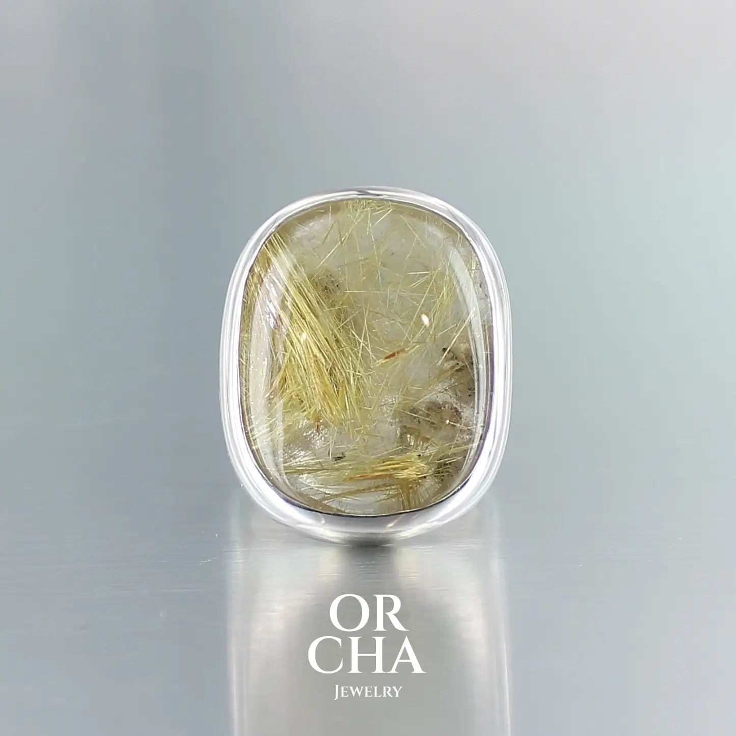 Grande bague pour femme en argent massif sertie d'un Quartz rutile doré à éclats d'hématites. Pierre cabochon en de forme rectangulaire, pierre naturelle incolore à inclusions dorés clairs . Pierre fine de qualité transparente. Taille 56. Bijoux unique, éthique et durable présenté dans un coffret de notre marque, prêt à offrir. Livraison offerte, certificat d'authenticité, service client.