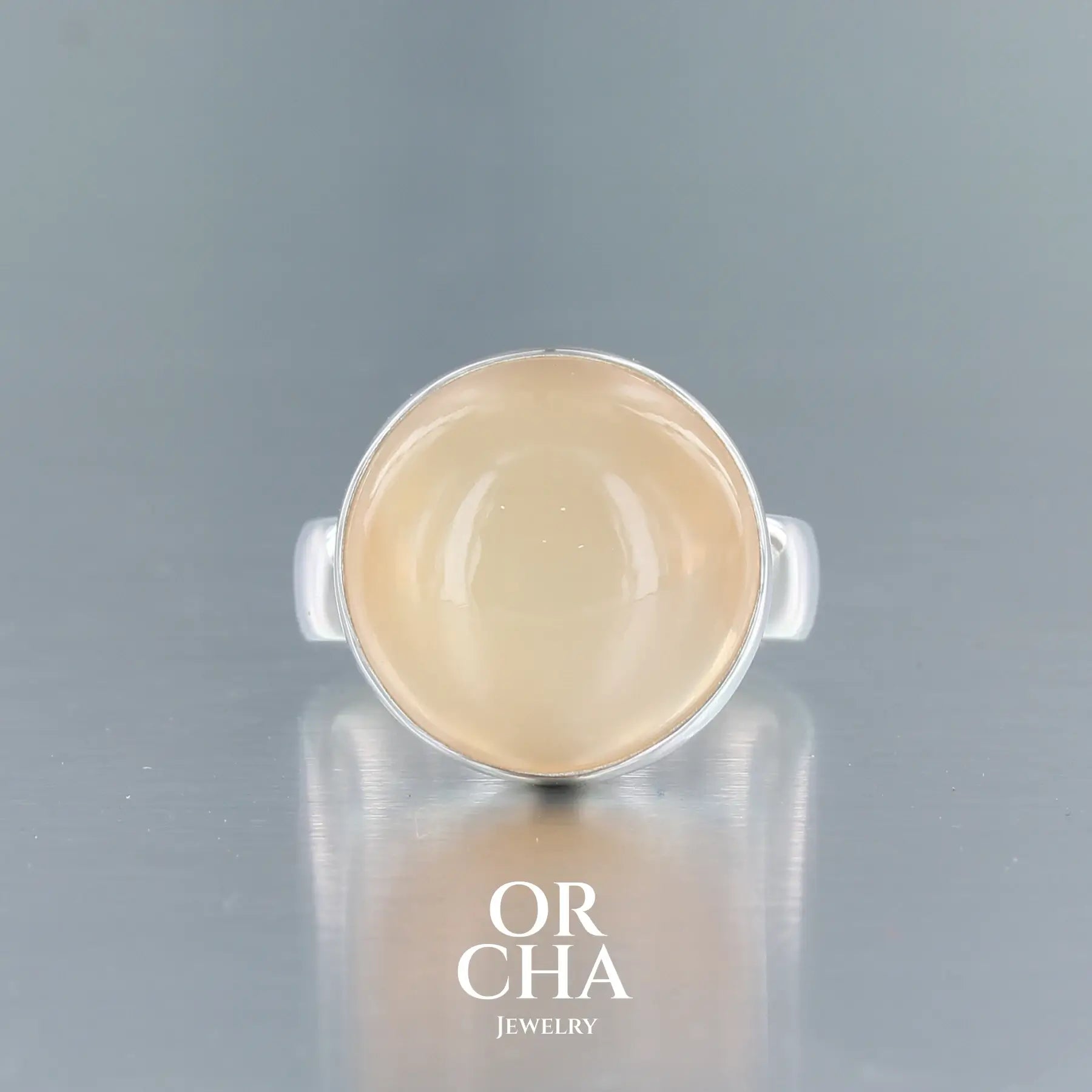 Bague contemporaine en argent massif pour femme sertie d'une pierre de lune orange. Pierre cabochon de forme ronde. Pierre transparente naturelle de couleur orange à chatoyance et effet satiné. Bague réalisée en taille 56. Bijoux unique, éthique et durable présenté dans un coffret de notre marque, prêt à offrir. Livraison offerte, certificat d'authenticité, service client.