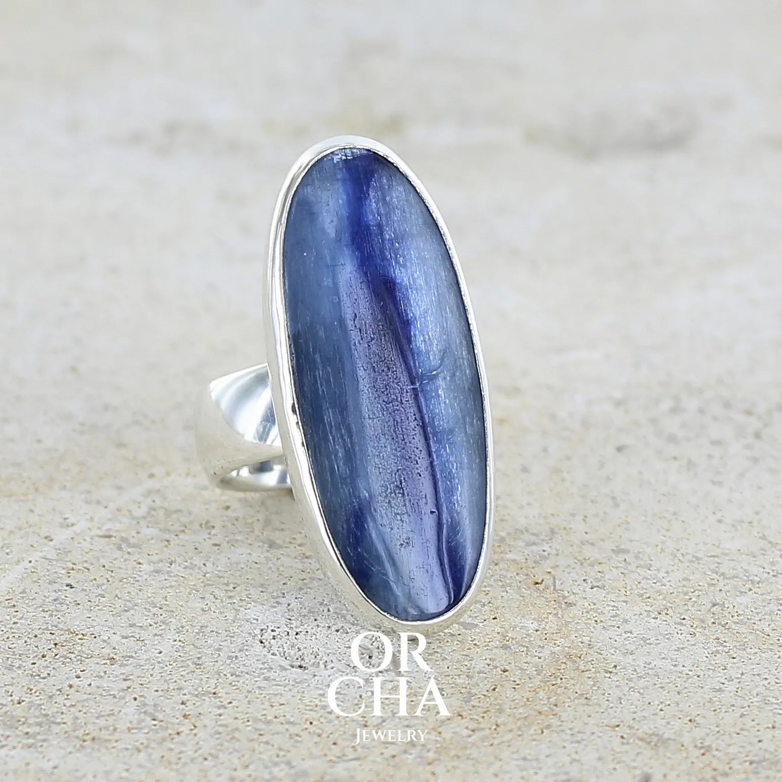 Bague pour femme en argent massif sertie d'un long cabochon ovale de cyanite bicolore de couleur bleu jean nacré. Pierre translucide à inclusions. Bijou unique réalisée en taille 54 par Orchajewelry