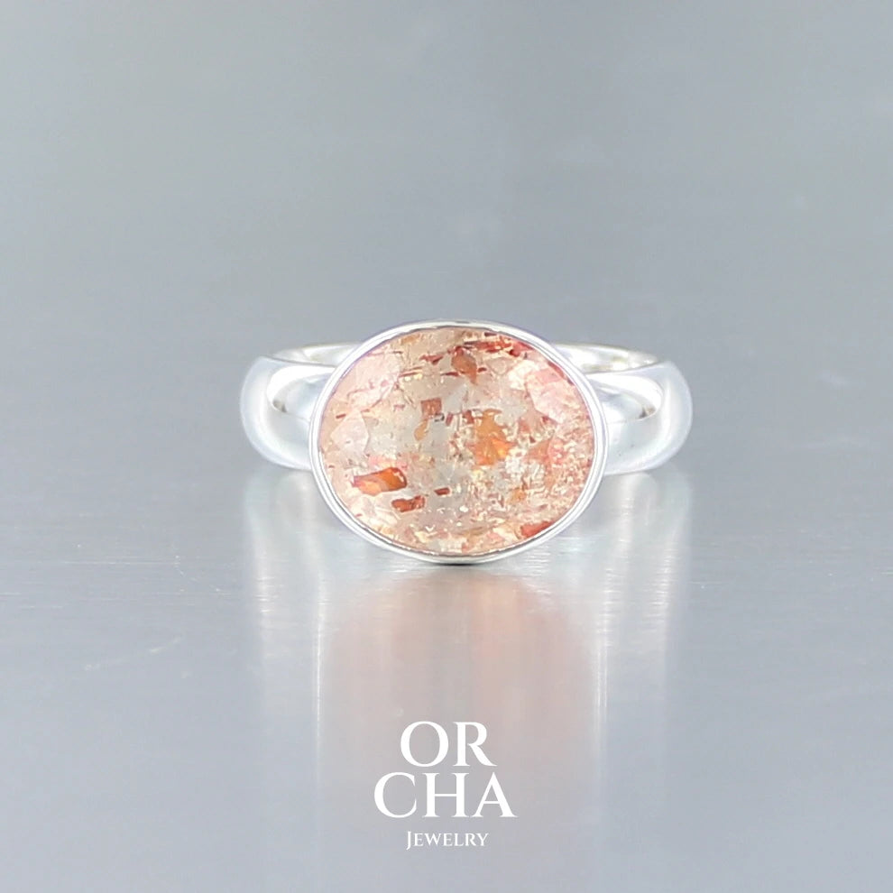Bague cocktail pour femme en argent massif sertie d'une Pierre de soleil. Pierre ovale facettée de de couleur orange clair avec des inclusions scintillantes d'hématites. Pierre naturelle. Bijoux unique, réalisé en taille 60, éthique et durable présenté dans un coffret de notre marque, prêt à offrir. Livraison offerte, certificat d'authenticité, service client.