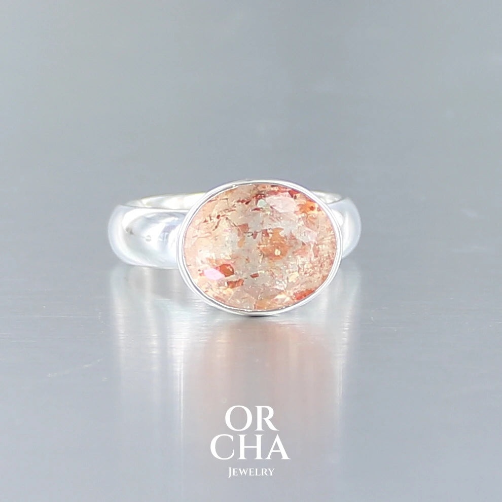 Bague cocktail pour femme en argent massif sertie d'une Pierre de soleil. Pierre ovale facettée de de couleur orange clair avec des inclusions scintillantes d'hématites. Pierre naturelle. Bijoux unique, réalisé en taille 60, éthique et durable présenté dans un coffret de notre marque, prêt à offrir. Livraison offerte, certificat d'authenticité, service client.
