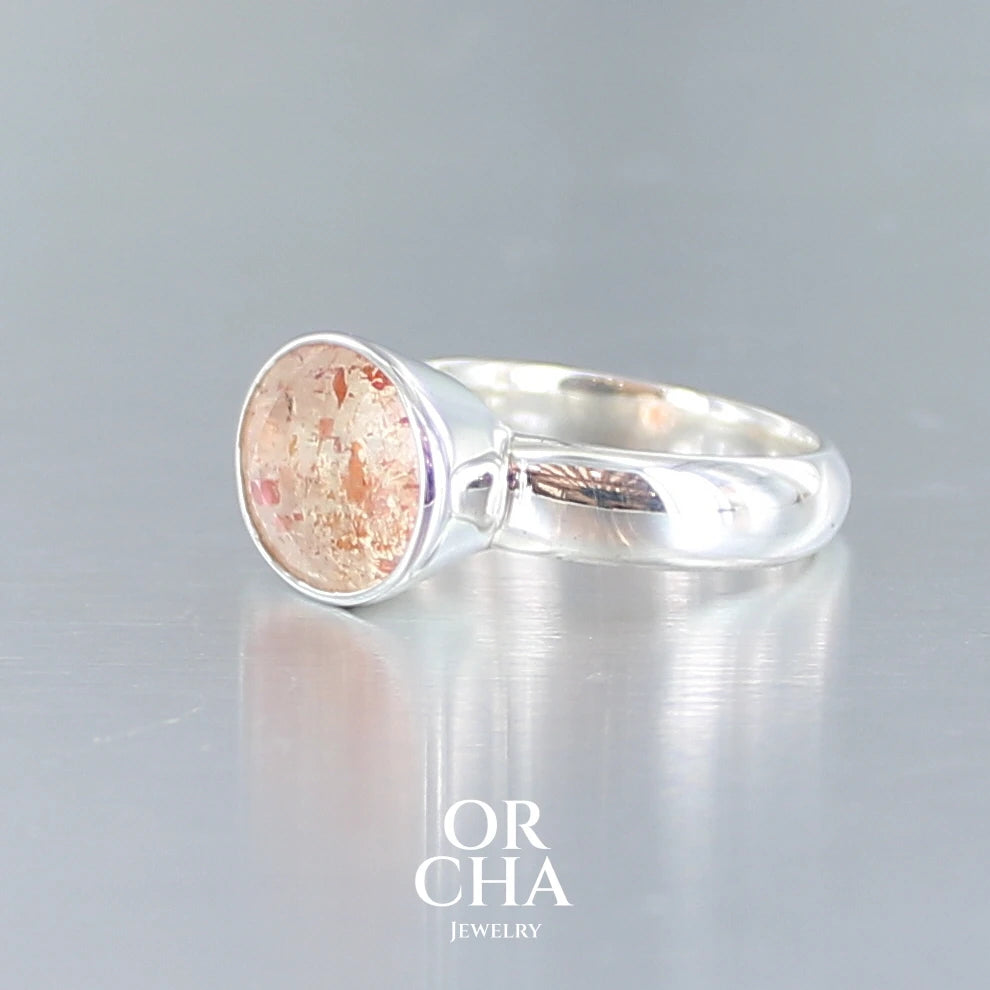 Bague cocktail pour femme en argent massif sertie d'une Pierre de soleil. Pierre ovale facettée de de couleur orange clair avec des inclusions scintillantes d'hématites. Pierre naturelle. Bijoux unique, réalisé en taille 60, éthique et durable présenté dans un coffret de notre marque, prêt à offrir. Livraison offerte, certificat d'authenticité, service client.