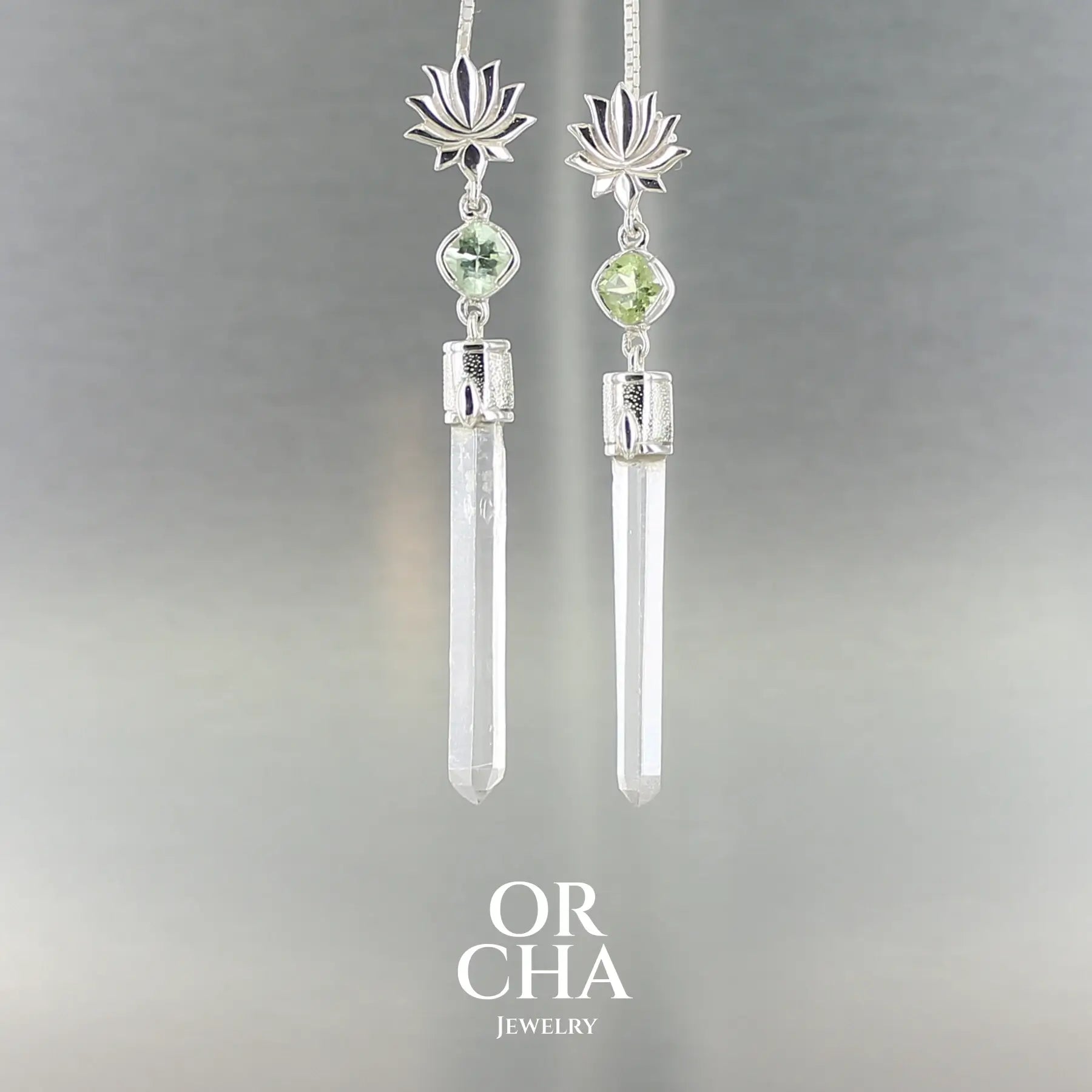 Paire de boucles d’oreilles pendantes en argent massif, ornées d’un motif de lotus stylisé en haut du lobe. Sous le lotus, une petite gemme taillée de couleur vert d’eau — une tourmaline verte — est sertie de manière délicate. Elle est suivie d’un cristal de roche naturel, long et fin, transparent, non taillé, en forme de pointe verticale. L’ensemble forme une silhouette élancée, à la fois pure et élégante.