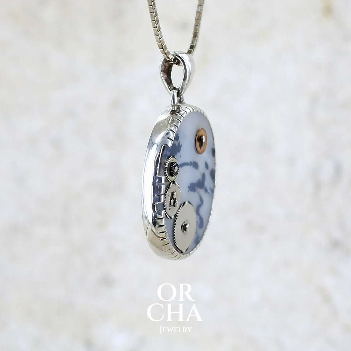 Détail du sertissage d'un pendentif steam punk pour homme en argent massif sertie d'une tranche ovale d'agate dendritique de couleur gris et noir, orné de mécanismes de montre. Pierre translucide à inclusions de type tâches d'encre de dendrites de manganèse noires contrastées. Bijou unique réalisé par Orchajewelry