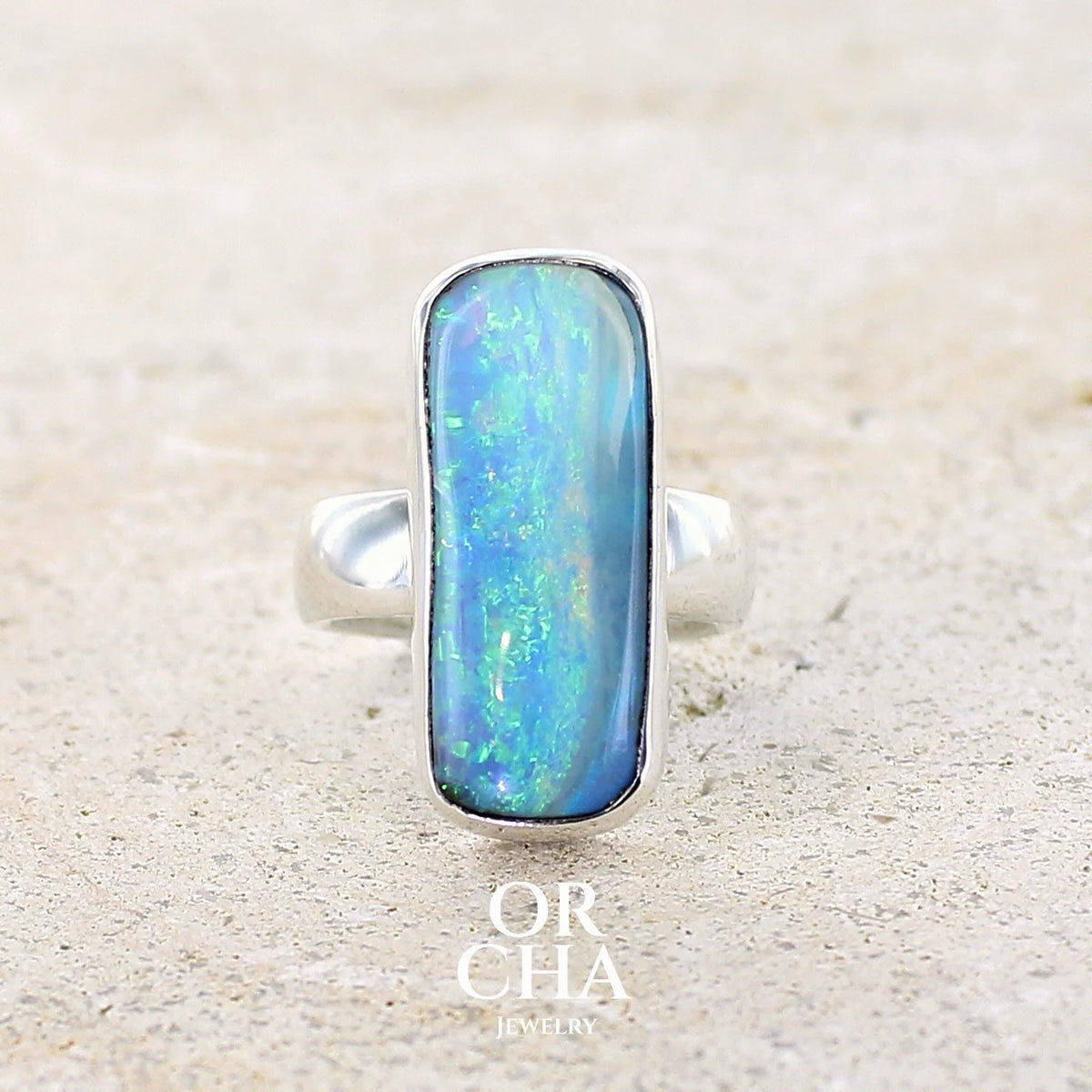 Bague en argent massif, avec une grande opale boulder rectangulaire sertie en cabochon. La pierre présente des reflets irisés bleu-vert intenses, ponctués de touches orangées. Le métal est lisse et poli, avec un anneau droit, épais et solide. Bague réalisée en taille 60. Bijoux unique par Orcha jewelry