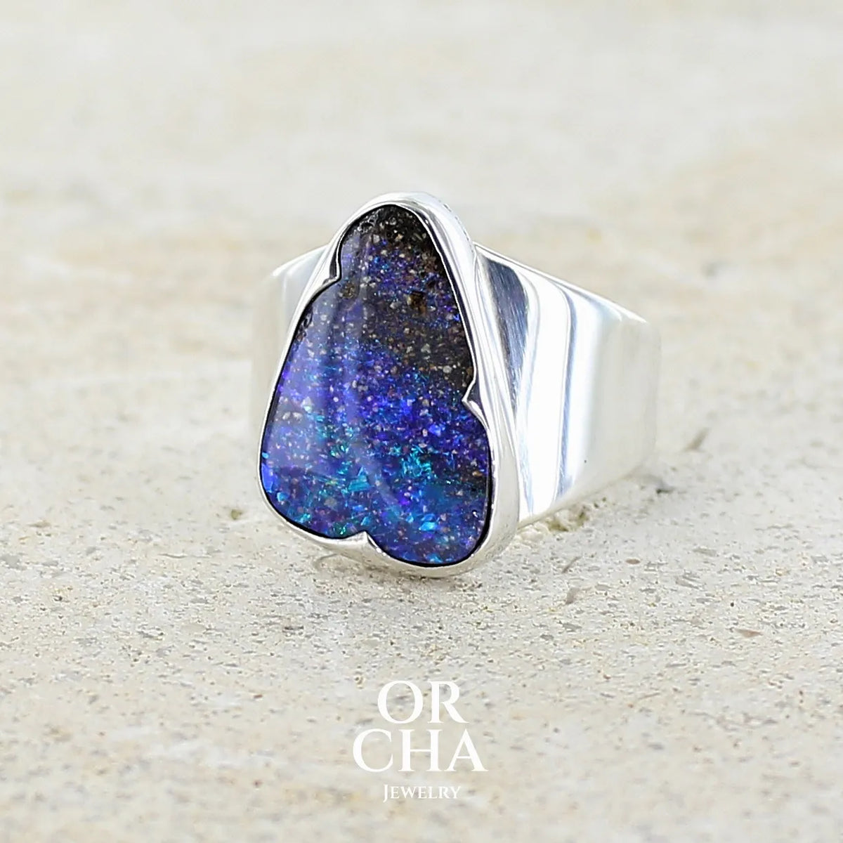 Profil gauche d'une bague pour femme en argent massif 925 sertie d'une opale boulder aux feux multicolores bleus violet et verts. Cabochon d'opale massive naturelle taillée en forme d'aile. Pierre de qualité opaque. Bijoux unique réalisé en taille 58 par Orcha Jewelry
