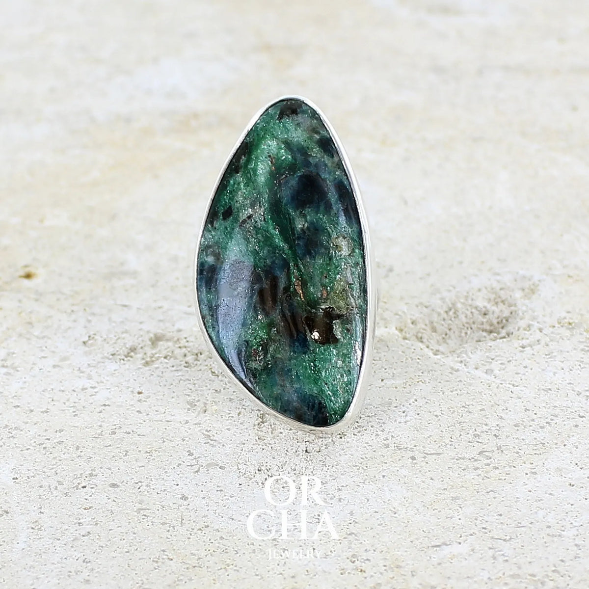 Bague pour femme en argent massif sertie d'un cabochon de pierre verte scintillante à inclusions bleues et noires. Pierre de qualité opaque et de forme libre. Bijou unique réalisée en taille 56 par Orchajewelry