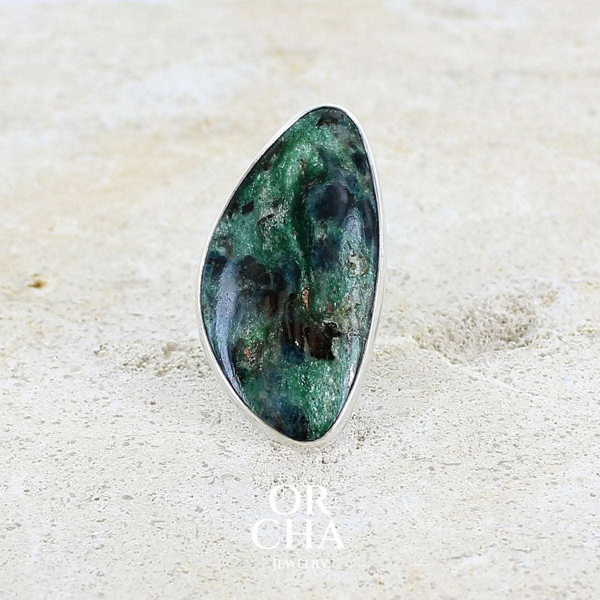 Bague pour femme en argent massif sertie d'un cabochon de pierre verte scintillante à inclusions bleues et noires. Pierre de qualité opaque et de forme libre. Bijou unique réalisée en taille 56 par Orchajewelry