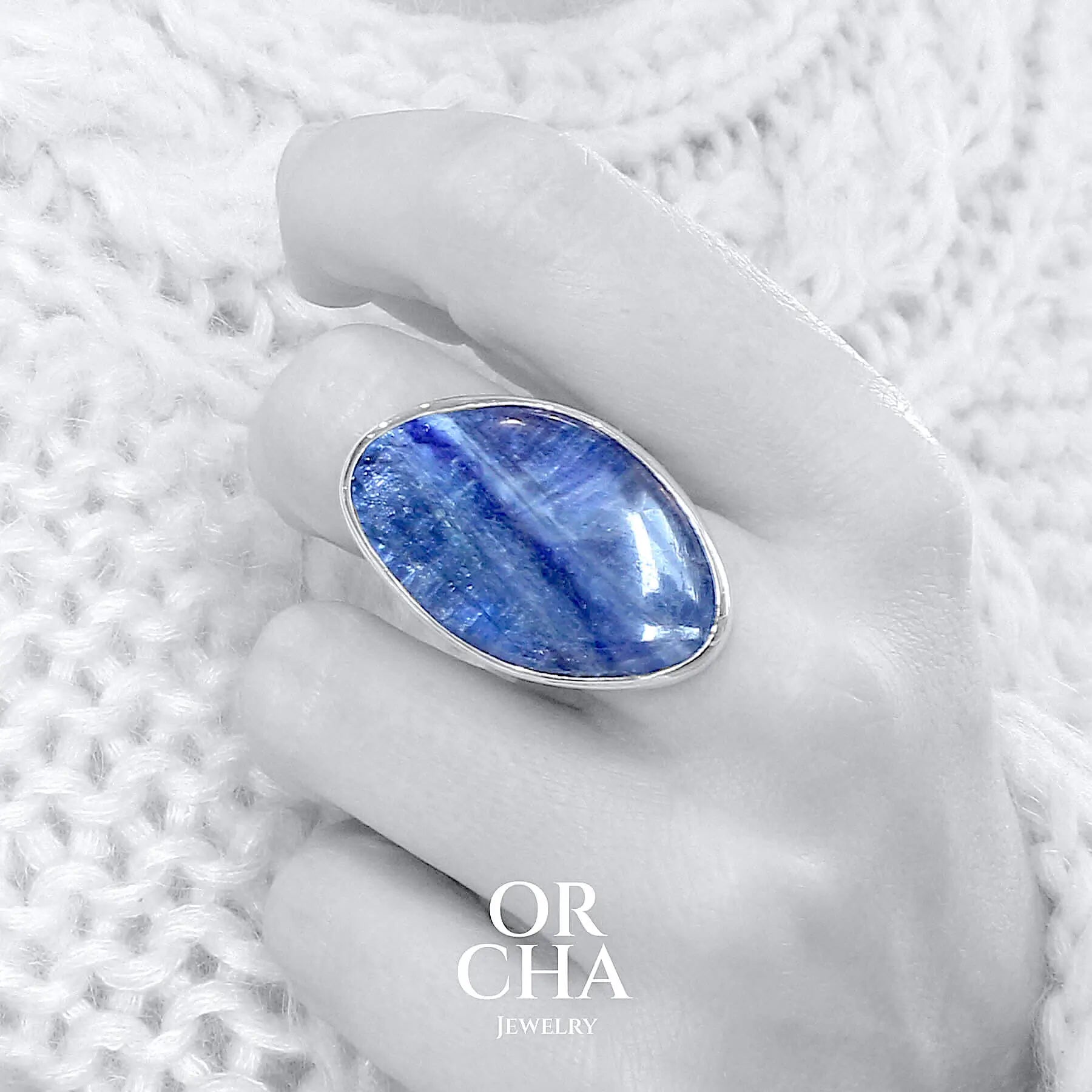 Bague portée sur une main de femme en argent massif sertie d'une Cyanite. Pierre cabochon de forme libre, pierre naturelle de couleur bleu denim avec des nuances de bleues. Qualité translucide. Taille 56. Bijoux unique, éthique et durable présenté dans un coffret de notre marque, prêt à offrir. Livraison offerte, certificat d'authenticité, service client.