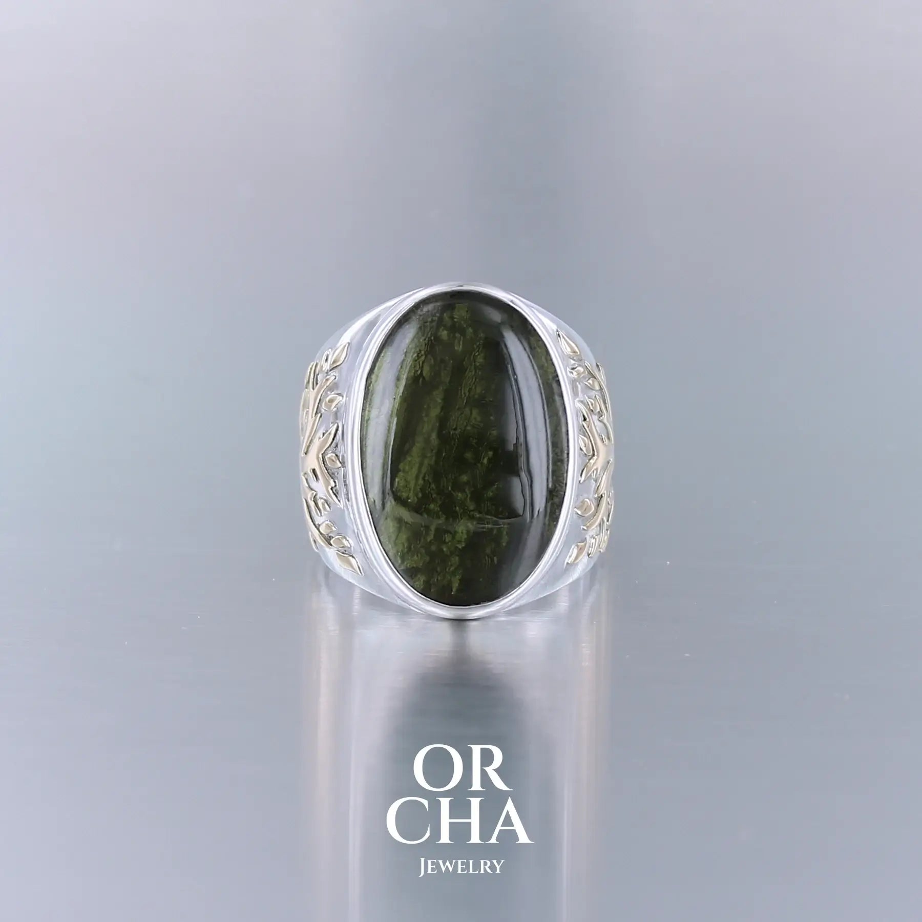 Bague pour homme en argent massif et or 18k où s'illustrent de chaque cotés de la bague des Arbres de vies en or. Cette bague est sertie d'un cabochon de moldavite de couleur vert foncé. Pierre naturelle transparente qui laisse voir le fond de la pierre laissé brute. Taille 64. Bijoux unique, éthique et durable présenté dans un coffret de notre marque, prêt à offrir. Livraison offerte, certificat d'authenticité, service client.