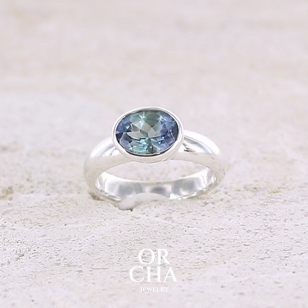 Bague solitaire pour femme en argent massif sertie d'une tanzanite bicolore. Pierre facettée de forme ovale de qualité transparente, propre à l'oeil. Pierre naturelle de couleur bleu verte et logés reflets violets. Bague réalisée en taille 50. Bijoux unique, éthique et durable présenté dans un coffret de notre marque, prêt à offrir. Livraison offerte, certificat d'authenticité, service client.