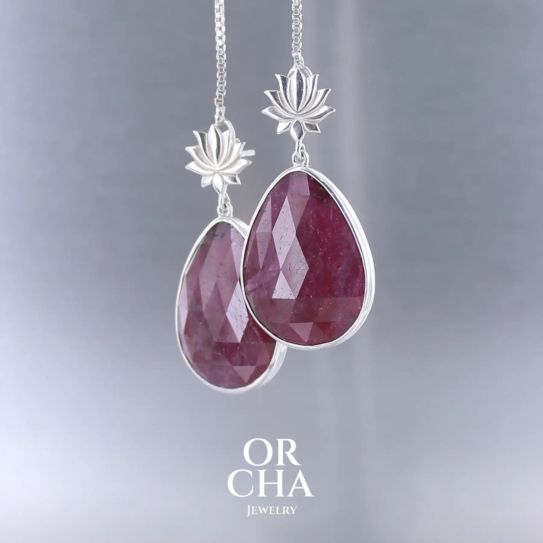 Boucles d'oreilles lotus pour femme en argent massif sertie de rubis. Cabochons facettés en forme de goutte de qualité opaque. Pierre naturelle de couleur rouge violacé. Le clou des boucles représente un lotus. Bijoux unique, éthique et durable présenté dans un coffret de notre marque, prêt à offrir. Livraison offerte, certificat d'authenticité, service client.