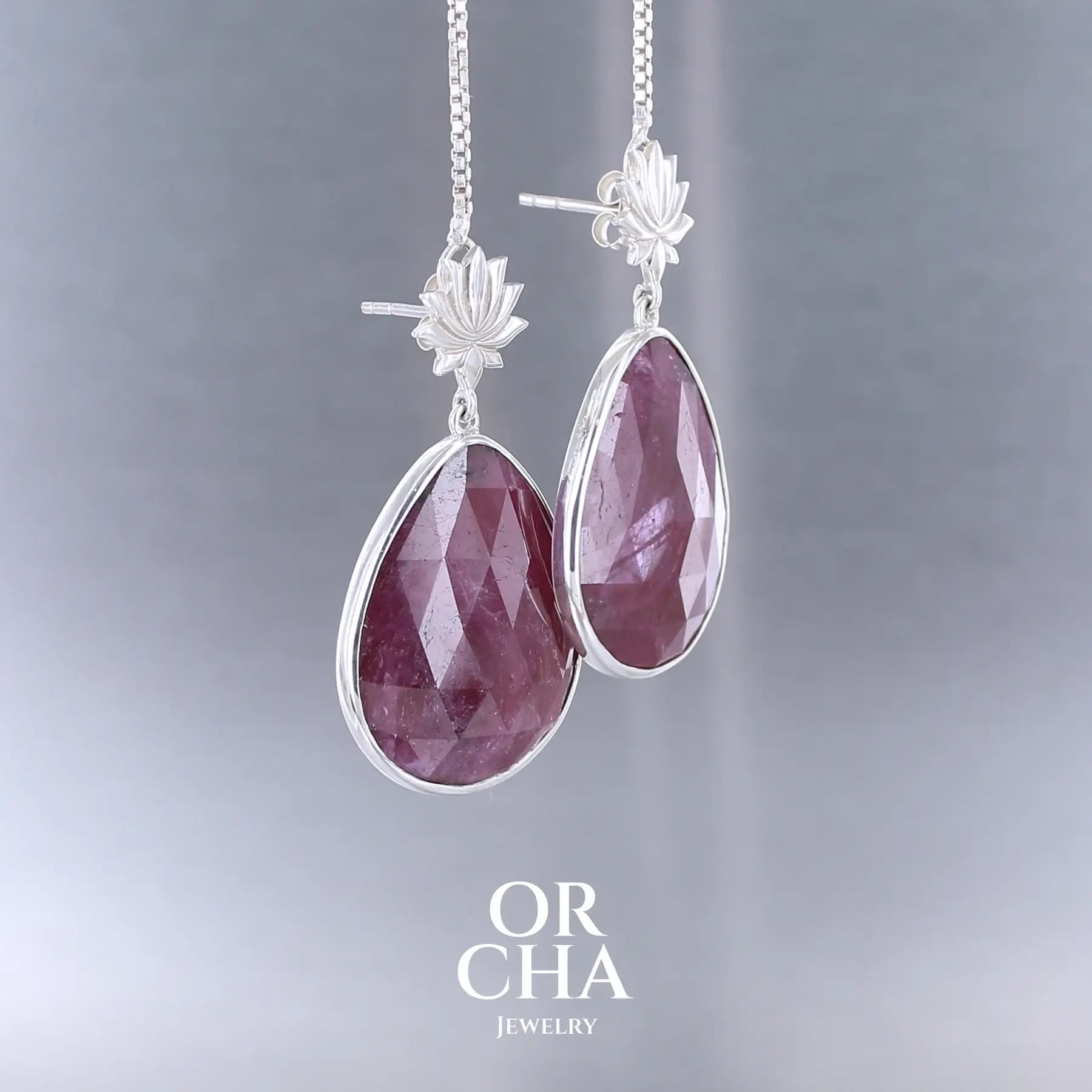 Boucles d'oreilles lotus pour femme en argent massif sertie de rubis. Cabochons facettés en forme de goutte de qualité opaque. Pierre naturelle de couleur rouge violacé. Le clou des boucles représente un lotus. Bijoux unique, éthique et durable présenté dans un coffret de notre marque, prêt à offrir. Livraison offerte, certificat d'authenticité, service client.