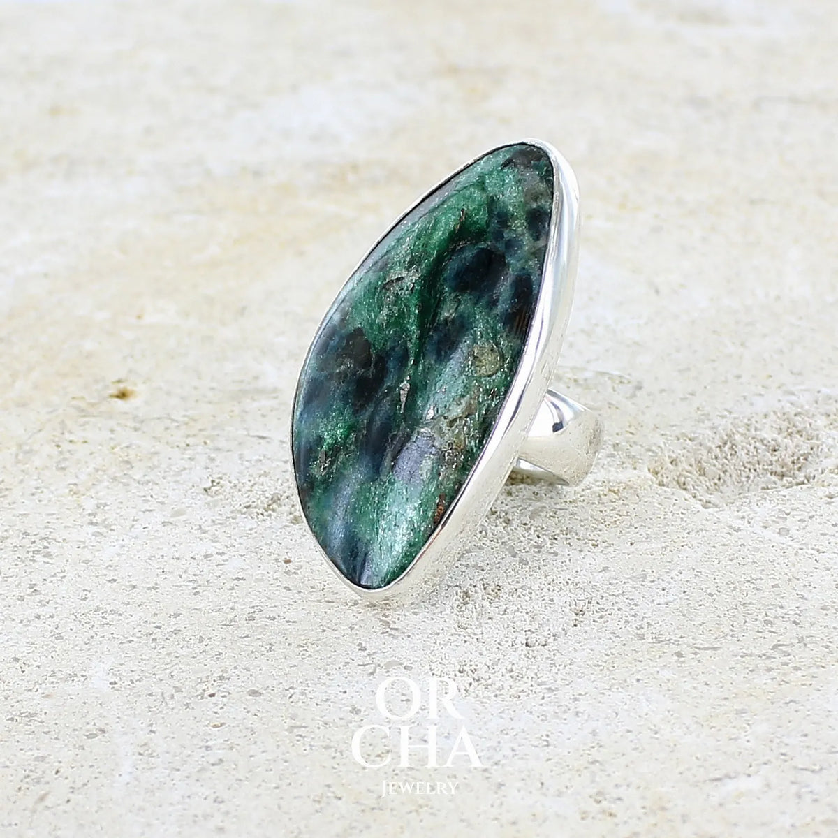 détail du sertissage d'une bague pour femme en argent massif sertie d'un cabochon de pierre verte scintillante à inclusions bleues et noires. Pierre de qualité opaque et de forme libre. Bijou unique réalisée en taille 56 par Orchajewelry
