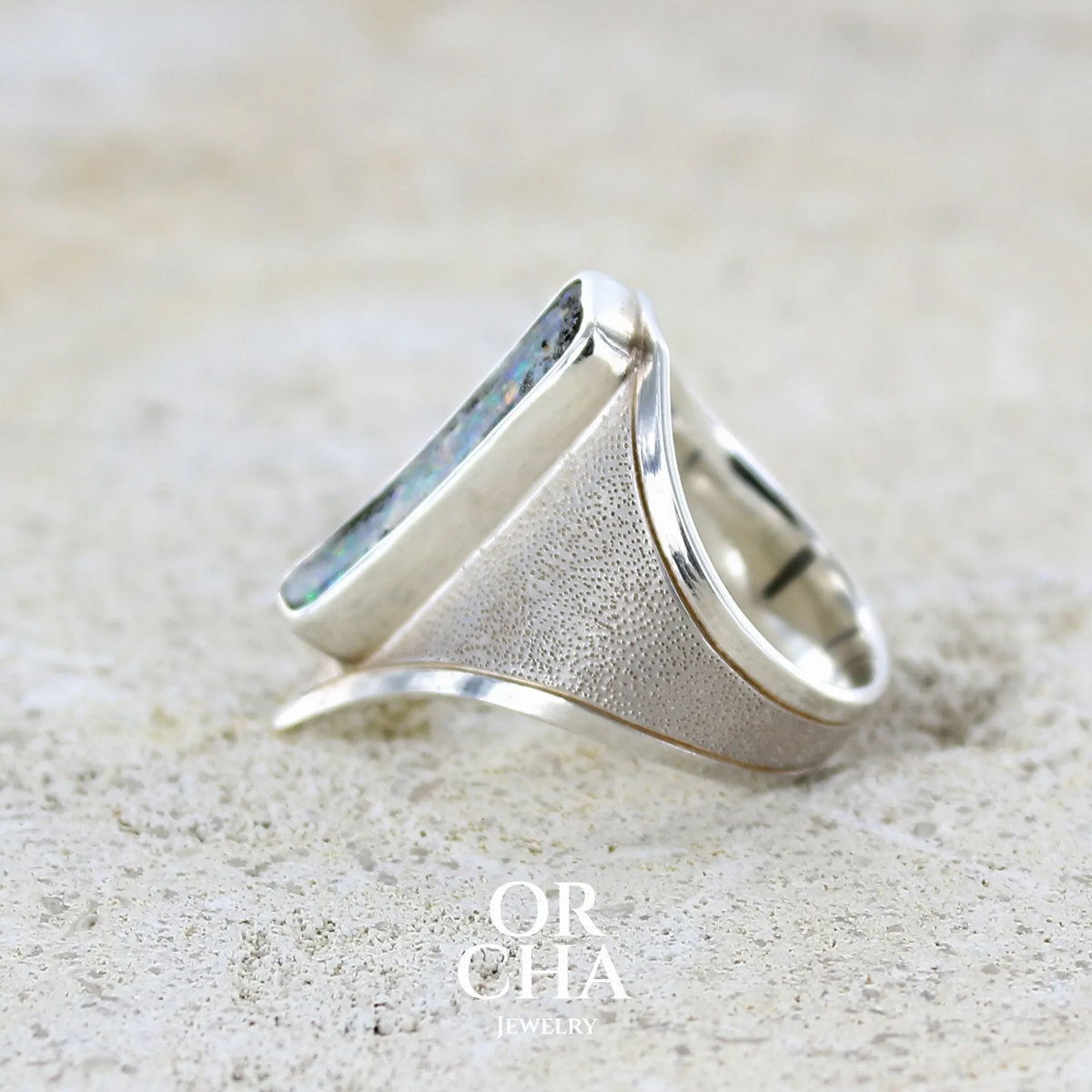 Détail d'une bague pour femme en argent massif 925 sertie d'une opale boulder aux feux multicolores. Cabochon d'opale massive naturelle taillée en forme de rectangle.. Pierre de qualité opaque. Bague unique réalisée en taille 55 par Orcha Jewelry.