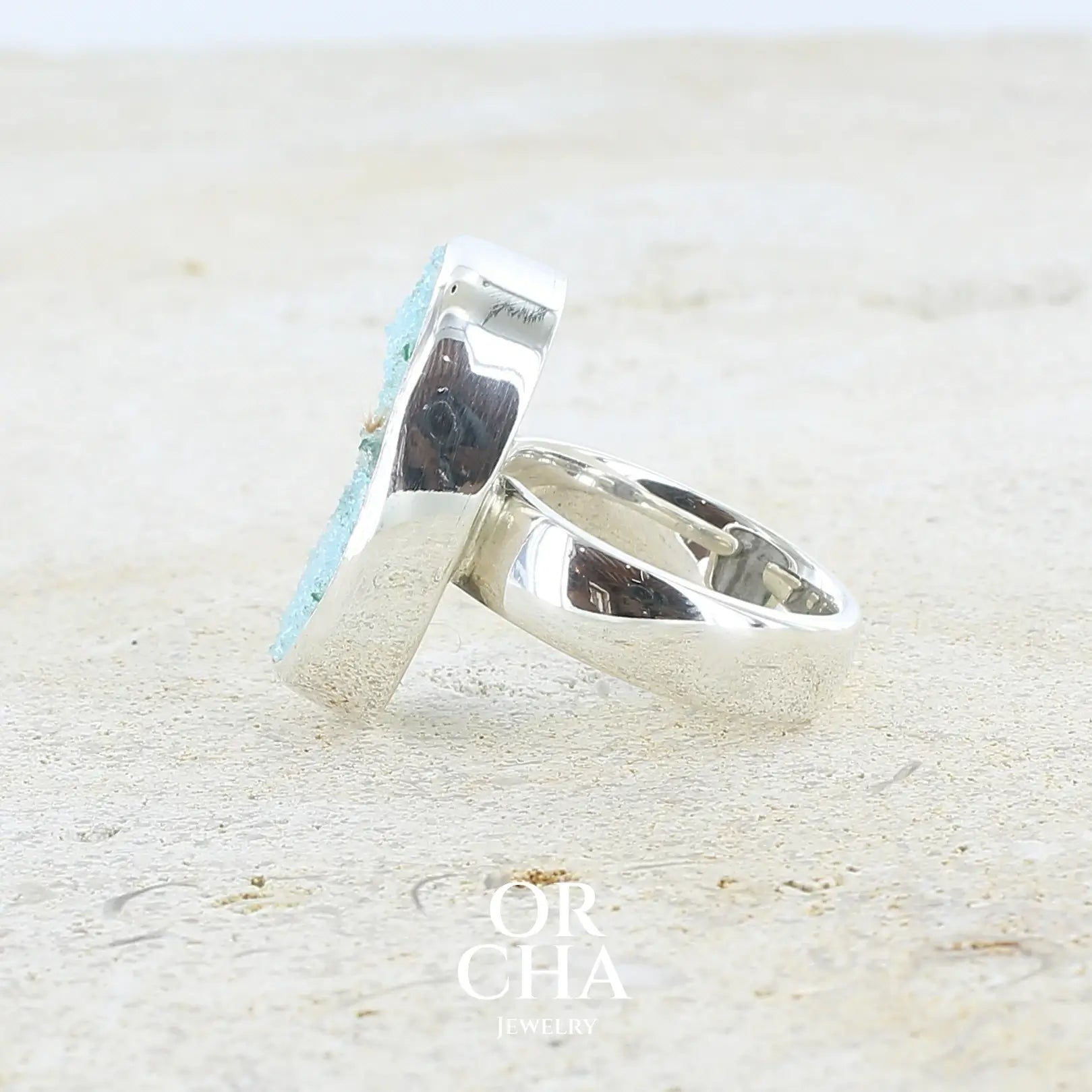 Détail de texture d'une bague pour femme en argent massif sertie d'un magnifique gemsilica druzy: chrysocale silicifiée recouverte de fins cristaux de quartz scintillants. Pierre de qualité opaque taillée en tranche de forme ovale. Pierre naturelle originaire de République dominicaine du Congo de couleur bleu clair turquoise. Bague réalisée en taille 58,5. Bijoux unique, éthique et durable présenté dans un coffret de notre marque, prêt à offrir. Livraison offerte, certificat d'authenticité, service client.
