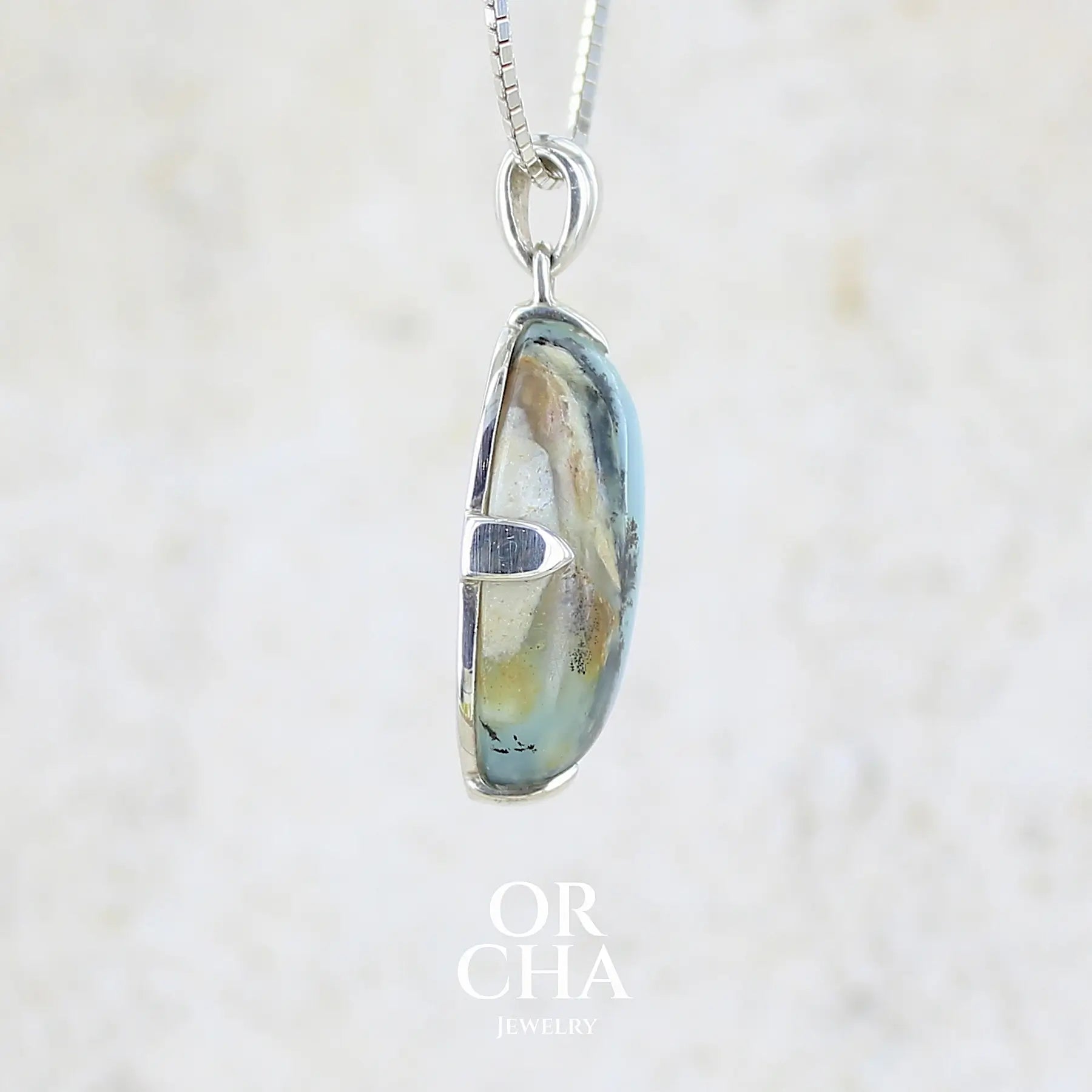 détail texture d'un pendentif pour femme et homme en argent massif, sertie d'un cabochon ovale d'opale bleue du Pérou de couleur bleu laiteux à inclusions de dendrites paysages noires et de roche mère marron. Pierre opaque et translucide. Bijoux unique, éthique et durable présenté dans un coffret de notre marque, prêt à offrir. Livraison offerte, certificat d'authenticité, service client.