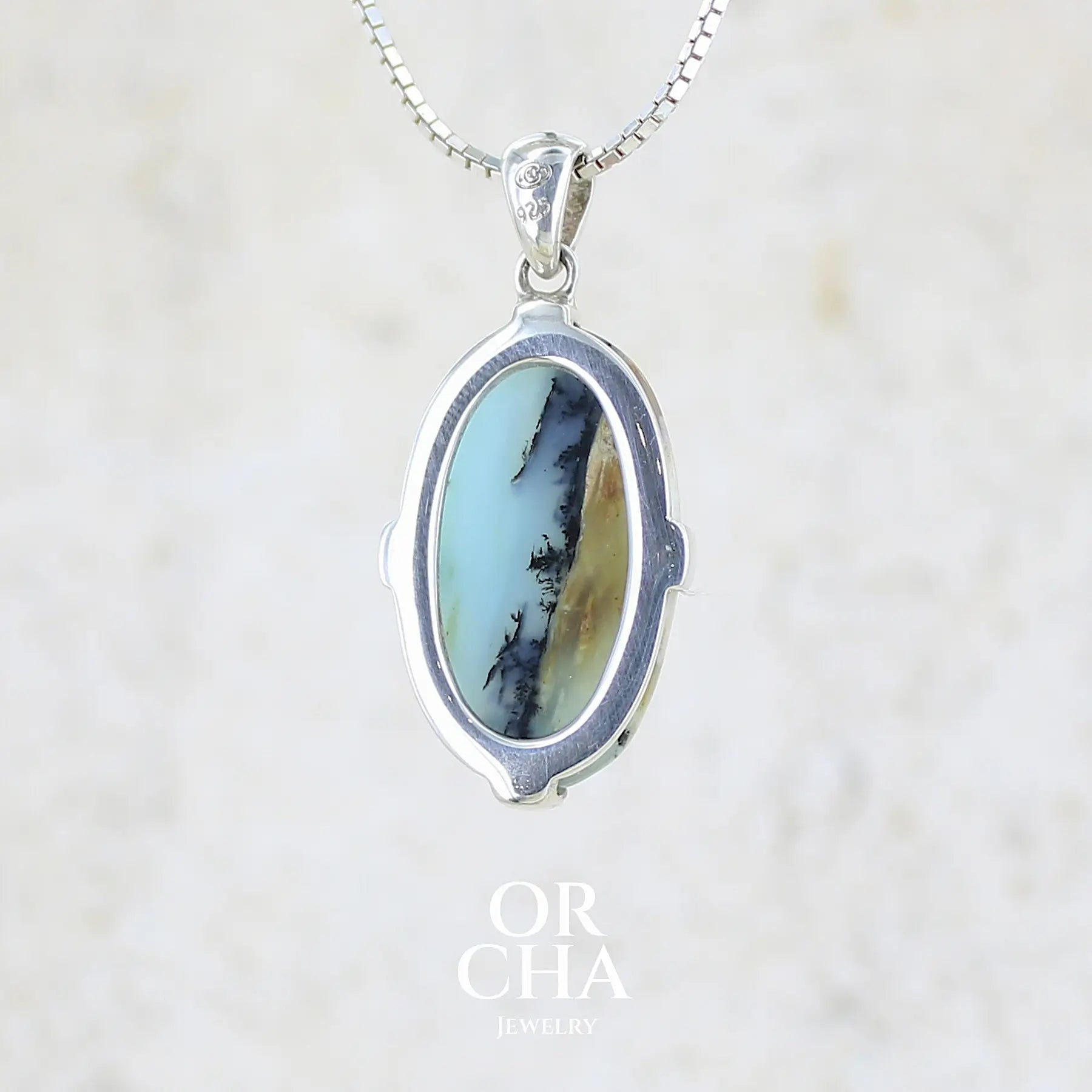 Vue de dos d'un pendentif pour femme et homme en argent massif, sertie d'un cabochon ovale d'opale bleue du Pérou de couleur bleu laiteux à inclusions de dendrites paysages noires et de roche mère marron. Pierre opaque et translucide. Bijoux unique, éthique et durable présenté dans un coffret de notre marque, prêt à offrir. Livraison offerte, certificat d'authenticité, service client.