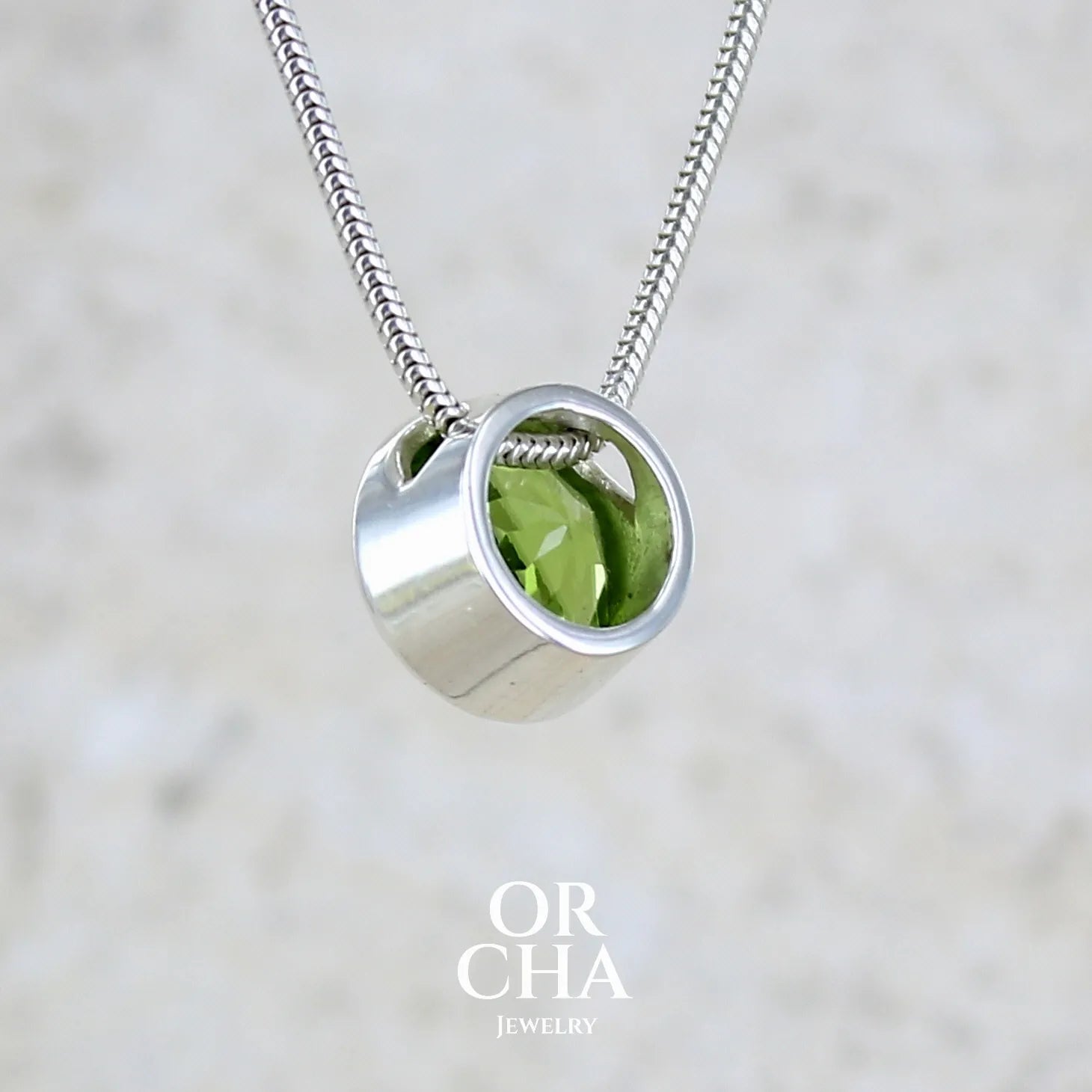 vue de dos d'un collier solitaire pour femme en argent massif serti d'un Péridot facettée de 4 carats. Pierre de forme ronde de qualité transparente, propre à l'oeil. Pierre naturelle de couleur vert olive doré intense et lumineuse. Pendentif monté sur une chaine en argent sterling de type maille serpent de 0,43 mm d'épaisseur et de 40 cm de longueur. Bijoux unique réalisé par Orchajewelry.
