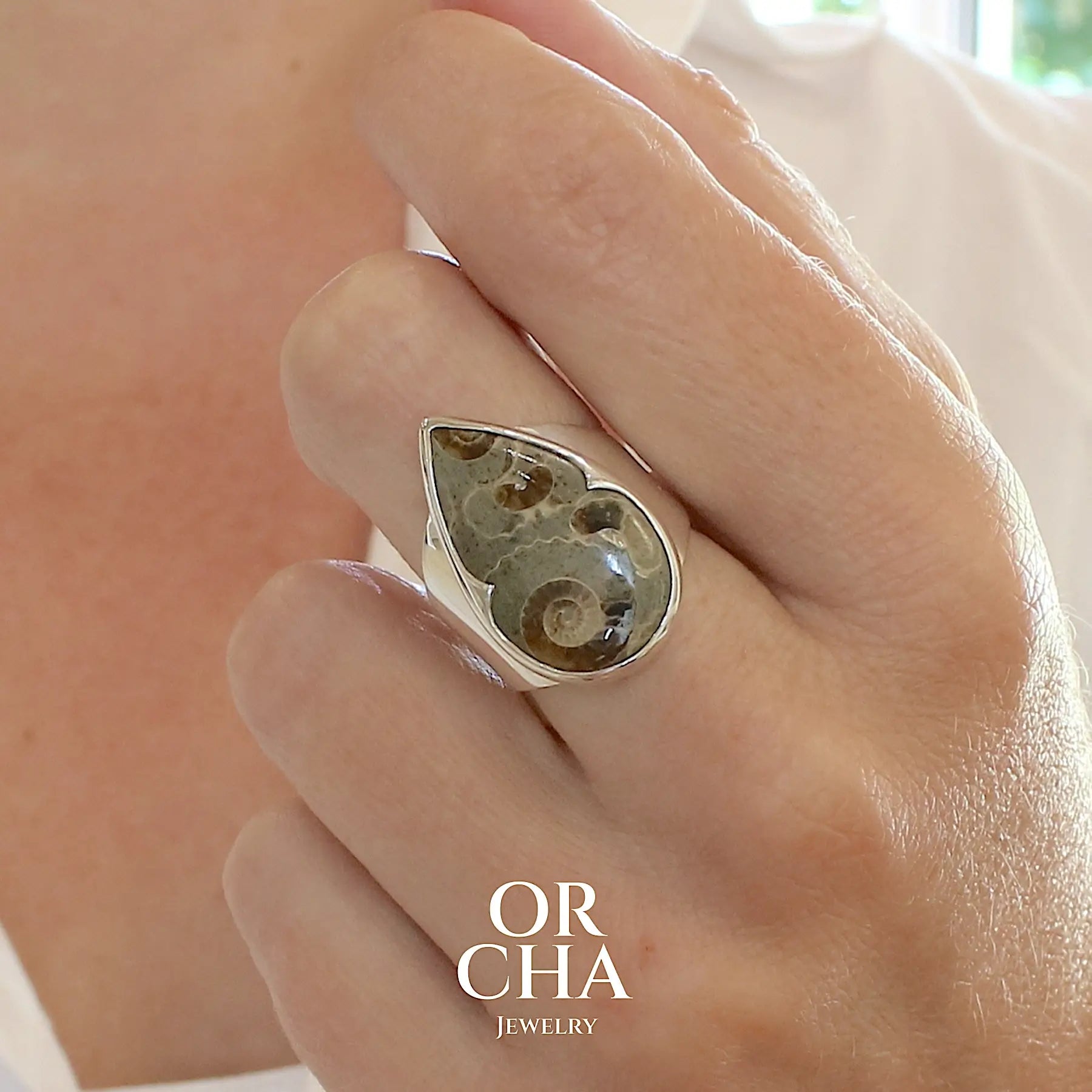 photo portée d'une bague pour femme en argent massif Bague en argent massif 925 sertie d'un marbre de Marston poire aux ammonites fossiles brun caramel sur fond gris taupe. Pierre fossile ornementale naturelle originaire Marston Magna, dans le Somerset en Angleterre. Bijoux uniques réalisé en taille 55 - Orcha jewelry
