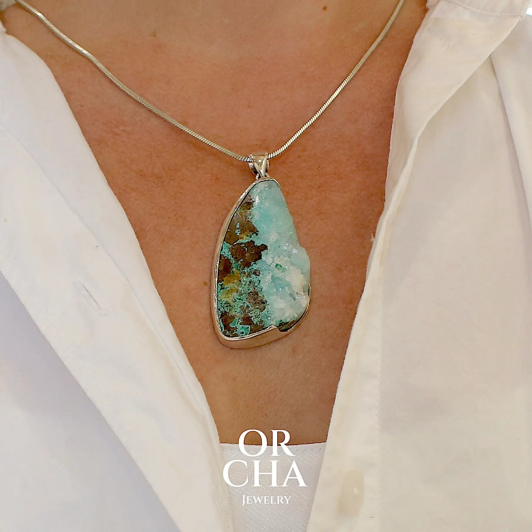 photo portée d'un pendentif en argent massif serti d'un magnifique gemsilica druze: chrysocolle silicifiée recouverte de fins cristaux de quartz scintillant. Pierre de qualité opaque brute en forme de goutte. Pierre naturelle originaire du Mexique de couleur bleue ciel turquoise claire.