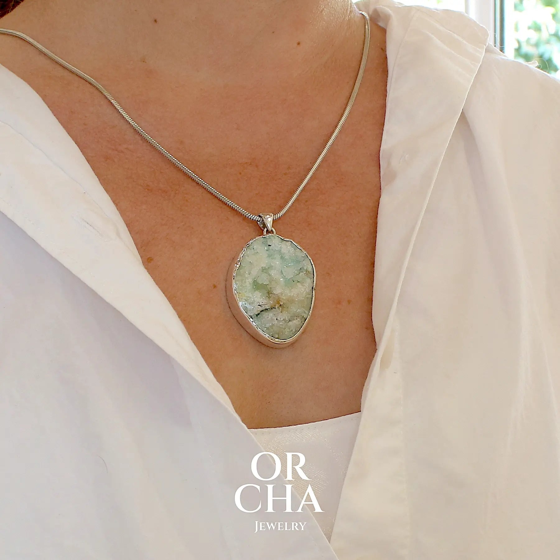 photo portée d'un pendentif en argent massif serti d'un magnifique gemsilica druze: chrysocolle silicifiée recouverte de fins cristaux de quartz scintillant. Pierre de qualité opaque brute de forme ovale. Pierre naturelle originaire du Mexique de couleur verte bleutée claire.