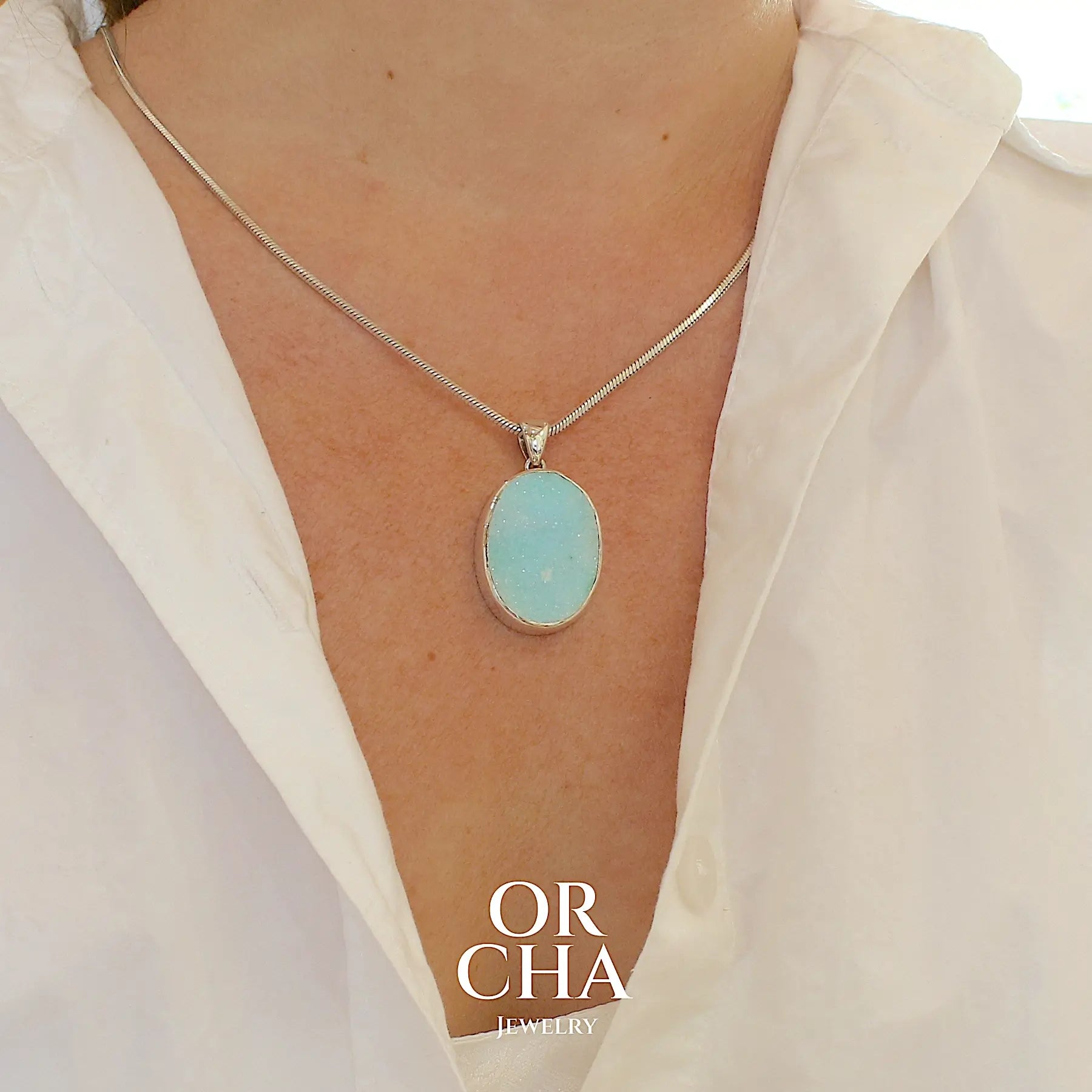photo sur modele d'un pendentif pour femme en argent massif sertie d'un magnifique gemsilica druzy, chrysocolle silicifiée recouverte de fins cristaux de quartz scintillant. Pierre de qualité opaque taillée en tranche de forme ovale. Pierre naturelle originaire de République dominicaine du Congo de couleur bleu clair turquoise. pierre rare et unique