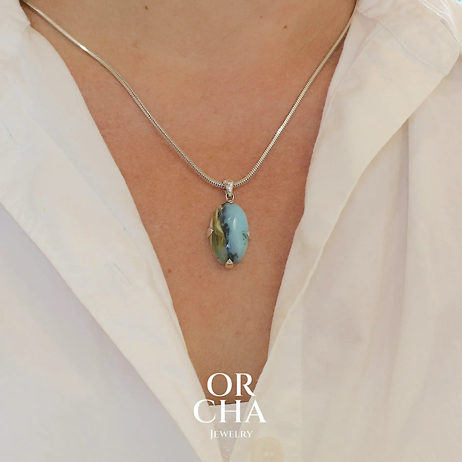 photo sur modele d'un pendentif pour femme et homme en argent massif, sertie d'un cabochon ovale d'opale bleue du Pérou de couleur bleu laiteux à inclusions de dendrites paysages noires et de roche mère marron. Pierre opaque et translucide. Bijoux unique, éthique et durable présenté dans un coffret de notre marque, prêt à offrir. Livraison offerte, certificat d'authenticité, service client.