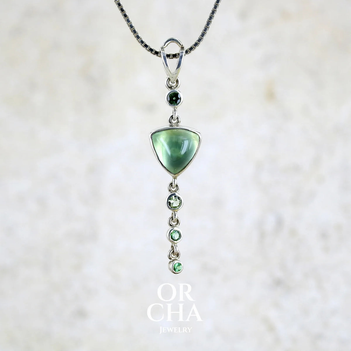 Pendentif pour femme en argent massif sertie d'un cabochon triangulaire de Préhnite ainsi que de petites tourmalines vertes formant une cascade colorée vert pastel lumineux. Pierre transparente à inclusions. Bijou unique réalisé par Orchajewelry