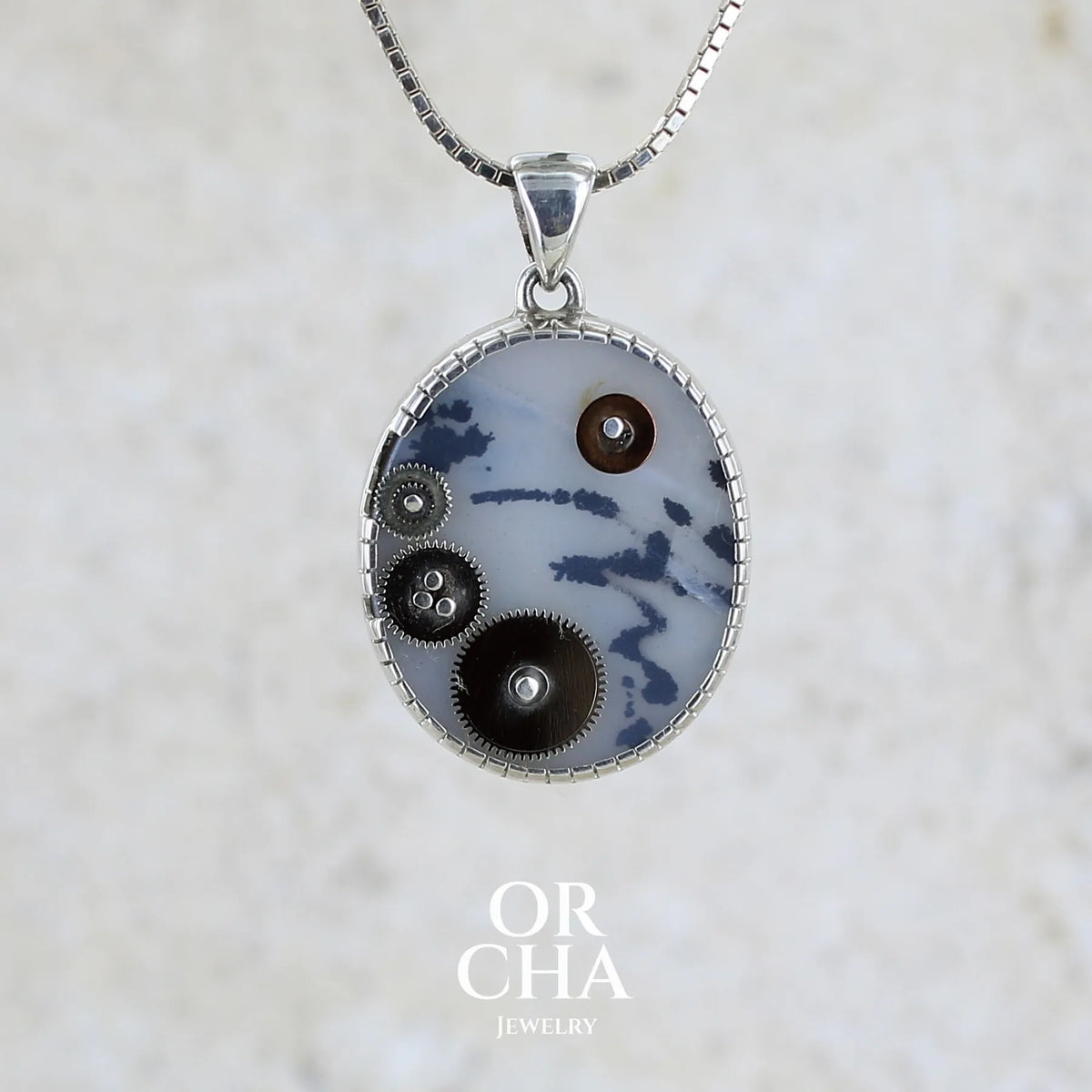 Pendentif steam punk pour homme en argent massif sertie d'une tranche ovale d'agate dendritique de couleur gris et noir, orné de mécanismes de montre. Pierre translucide à inclusions de type tâches d'encre de dendrites de manganèse noires contrastées. Bijou unique réalisé par Orchajewelry