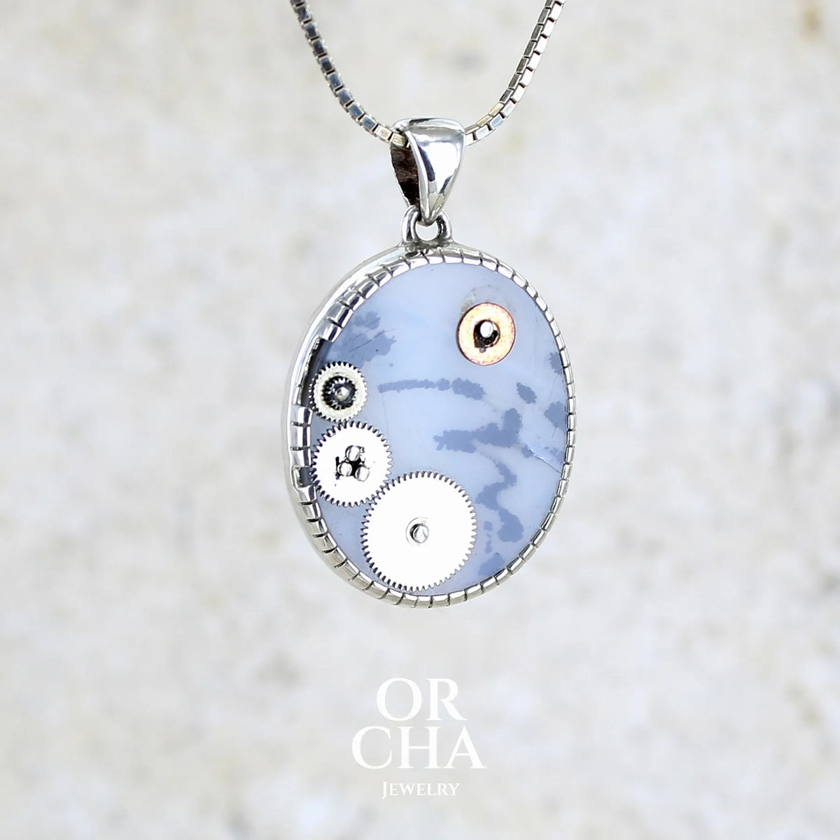 Pendentif steam punk pour homme en argent massif sertie d'une tranche ovale d'agate dendritique de couleur gris et noir, orné de mécanismes de montre. Pierre translucide à inclusions de type tâches d'encre de dendrites de manganèse noires contrastées. Bijou unique réalisé par Orchajewelry
