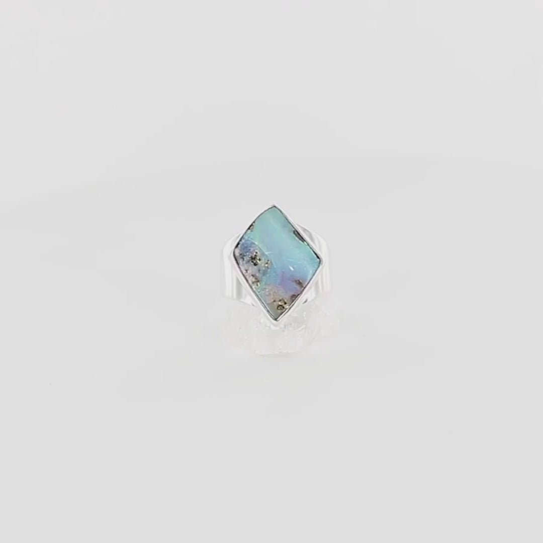 Bague en argent massif 925 sertie d'une opale boulder australienne de forme diamant asymétrique. La pierre présente une magnifique iridescence bleu-vert dominante avec des reflets violacés et quelques inclusions dorées naturelles. La monture simple et élégante en argent poli encadre la pierre, mettant en valeur ses couleurs changeantes. Bague réalisée en taille 56. Bijoux unique, éthique et durable présenté dans un coffret de notre marque, prêt à offrir. Livraison offerte, certificat d'authenticité, service