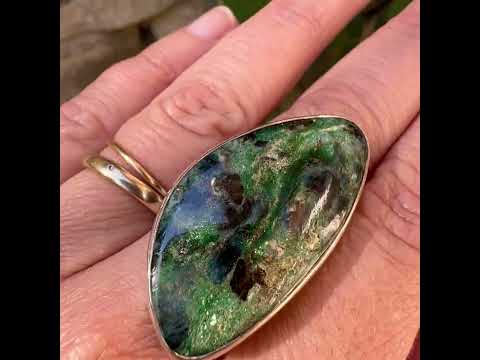 Bague pour femme en argent massif sertie d'un cabochon de pierre verte scintillante à inclusions bleues et noires. Pierre de qualité opaque et de forme libre. Bijou unique réalisée en taille 56 par Orchajewelry