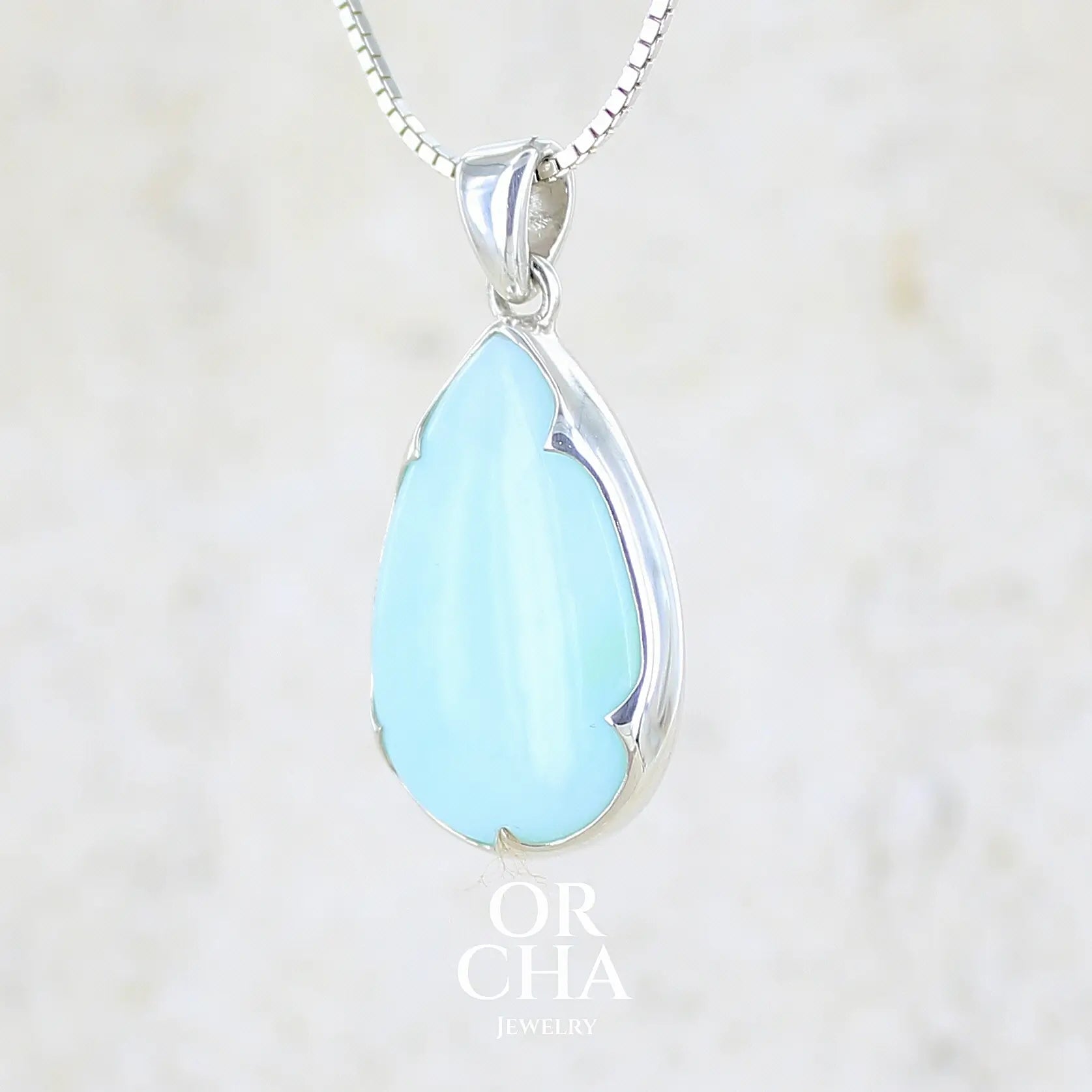 profil gauche d'un pendentif en argent massif, sertie d'un cabochon d'opale bleue du Pérou de couleur bleu ciel clair. Pierre en forme de goutte. Pierre opaque et translucide. Bijoux unique, éthique et durable présenté dans un coffret de notre marque, prêt à offrir. Livraison offerte, certificat d'authenticité, service client.