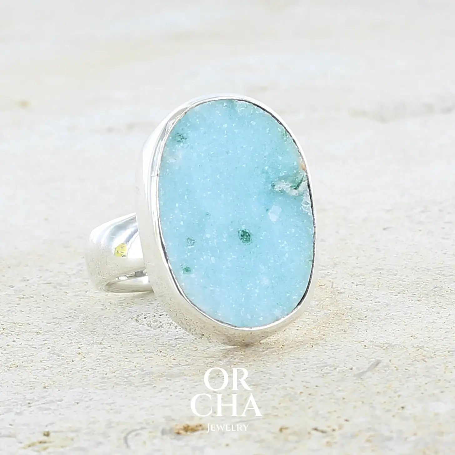 Profil droit d'une bague pour femme en argent massif sertie d'un magnifique gemsilica druzy: chrysocale silicifiée recouverte de fins cristaux de quartz scintillants. Pierre de qualité opaque taillée en tranche de forme ovale. Pierre naturelle originaire de République dominicaine du Congo de couleur bleu clair turquoise. Bague réalisée en taille 58,5. Bijoux unique, éthique et durable présenté dans un coffret de notre marque, prêt à offrir. Livraison offerte, certificat d'authenticité, service client.