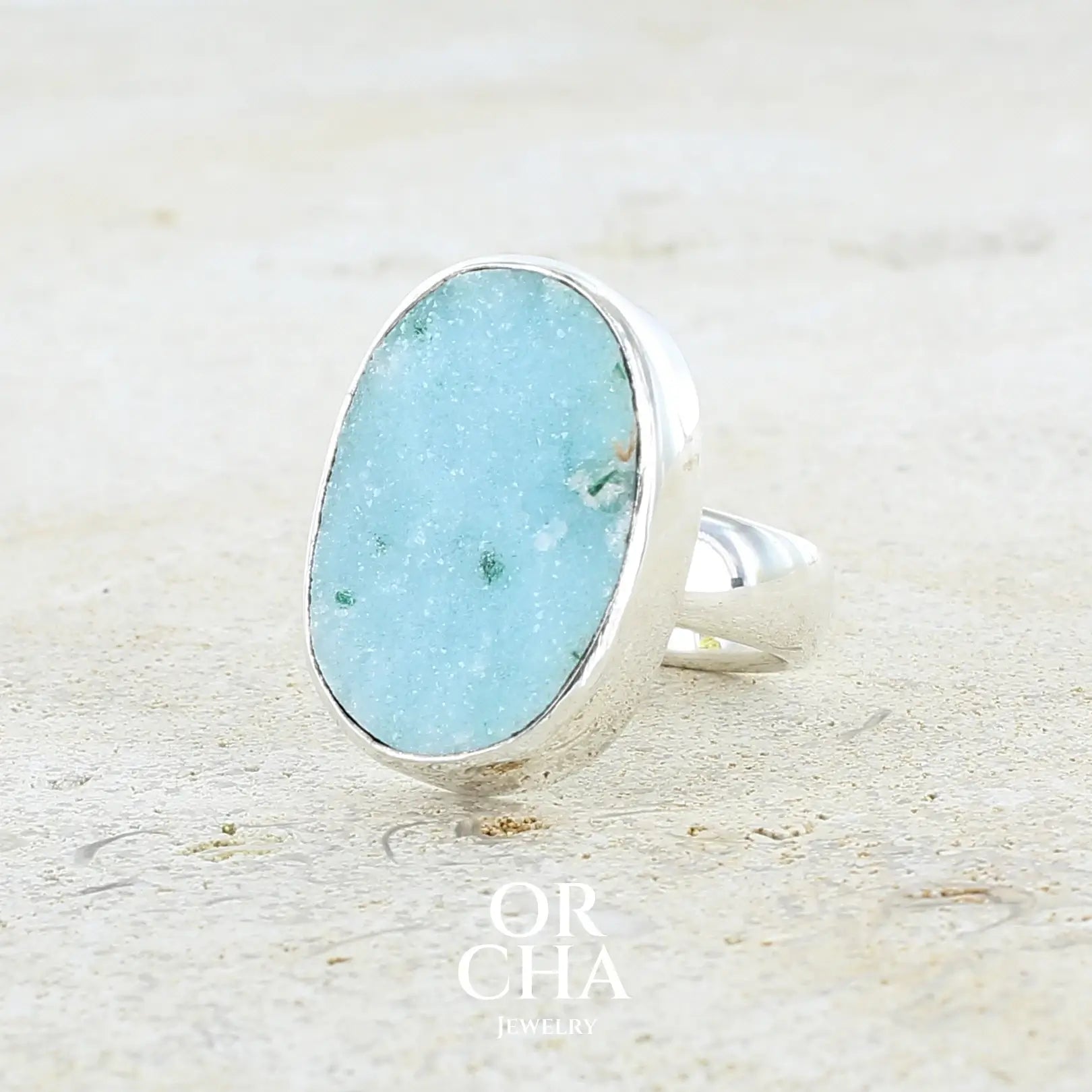 Profil gauche d'une bague pour femme en argent massif sertie d'un magnifique gemsilica druzy: chrysocale silicifiée recouverte de fins cristaux de quartz scintillants. Pierre de qualité opaque taillée en tranche de forme ovale. Pierre naturelle originaire de République dominicaine du Congo de couleur bleu clair turquoise. Bague réalisée en taille 58,5. Bijoux unique, éthique et durable présenté dans un coffret de notre marque, prêt à offrir. Livraison offerte, certificat d'authenticité, service client.