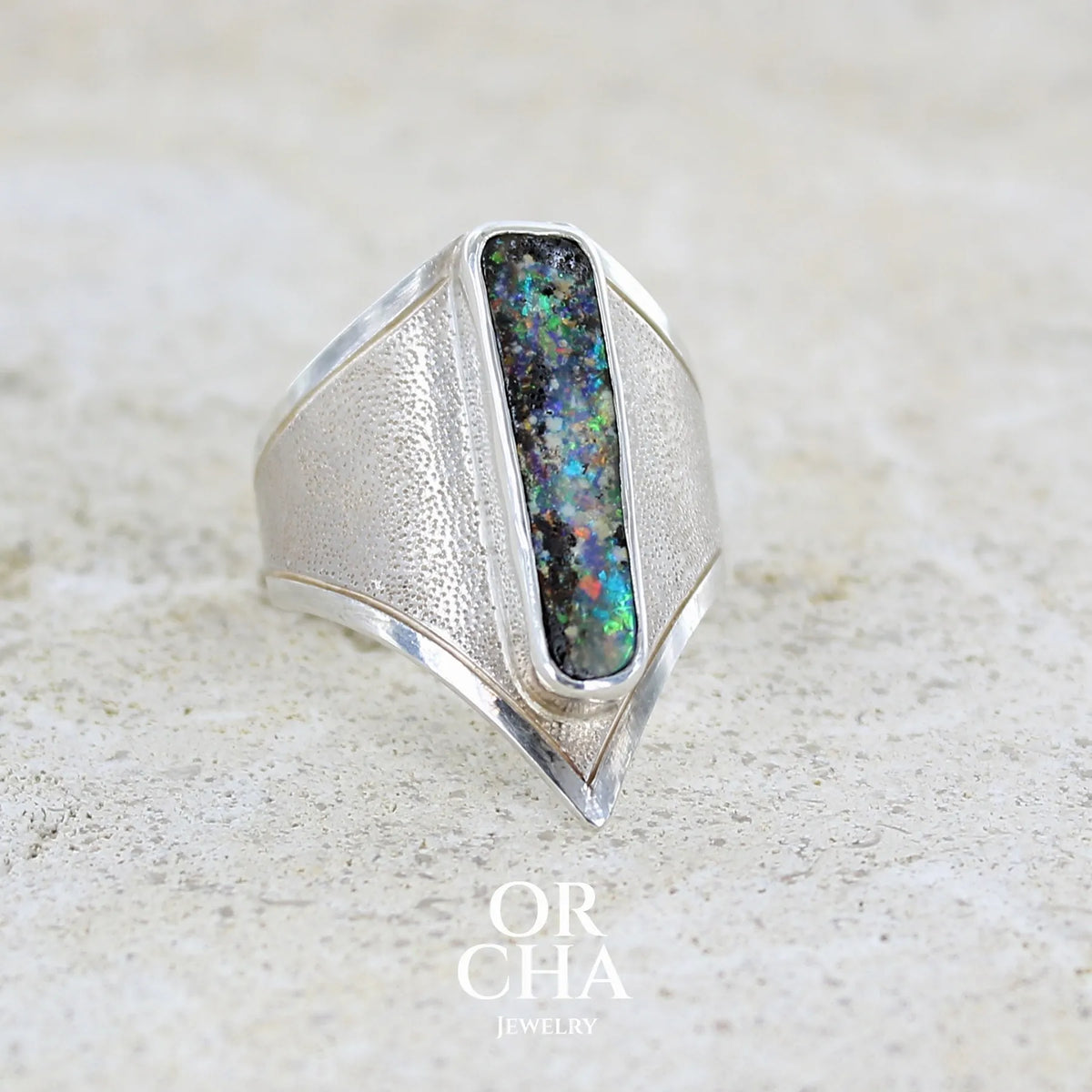 Profil droit d'une bague pour femme en argent massif 925 sertie d'une opale boulder aux feux multicolores. Cabochon d'opale massive naturelle taillée en forme de rectangle.. Pierre de qualité opaque. Bague unique réalisée en taille 55 par Orcha Jewelry.