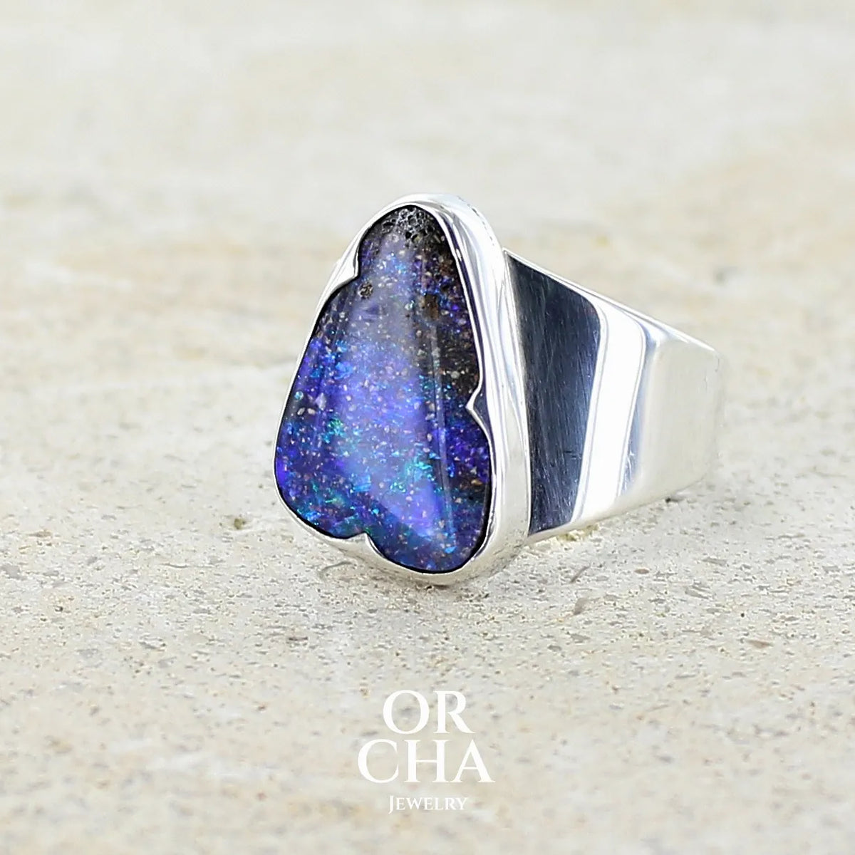 Profil gauche d'une bague pour femme en argent massif 925 sertie d'une opale boulder aux feux multicolores bleus violet et verts. Cabochon d'opale massive naturelle taillée en forme d'aile. Pierre de qualité opaque. Bijoux unique réalisé en taille 58 par Orcha Jewelry.