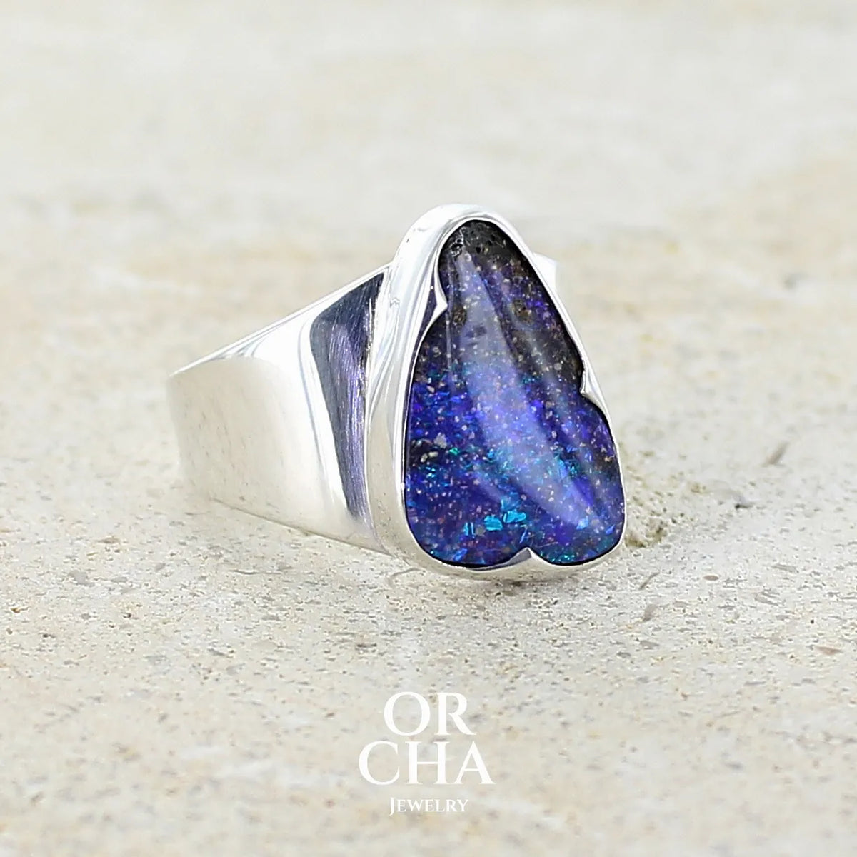 Profil gauche d'une bague pour femme en argent massif 925 sertie d'une opale boulder aux feux multicolores bleus violet et verts. Cabochon d'opale massive naturelle taillée en forme d'aile. Pierre de qualité opaque. Bijoux unique réalisé en taille 58 par Orcha Jewelry.