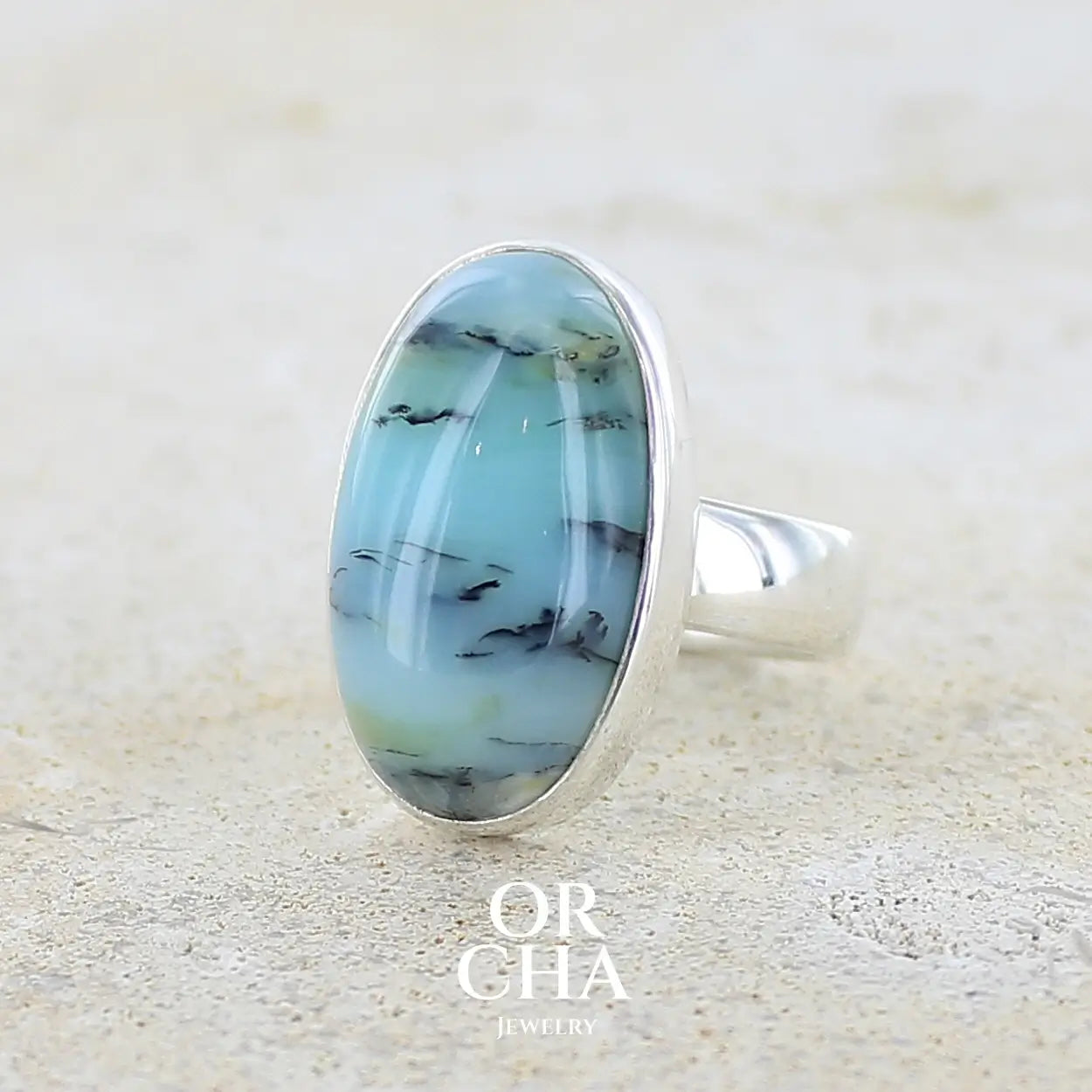Profil gauche d'une bague pour femme en argent massif, sertie d'un cabochon ovale d'opale bleue du Pérou de couleur bleu laiteux à inclusions de dendrites noires. Pierre naturelle de qualité opaque et translucide. Bijoux unique réalisé en taille 54,5 - OrchaJewelry