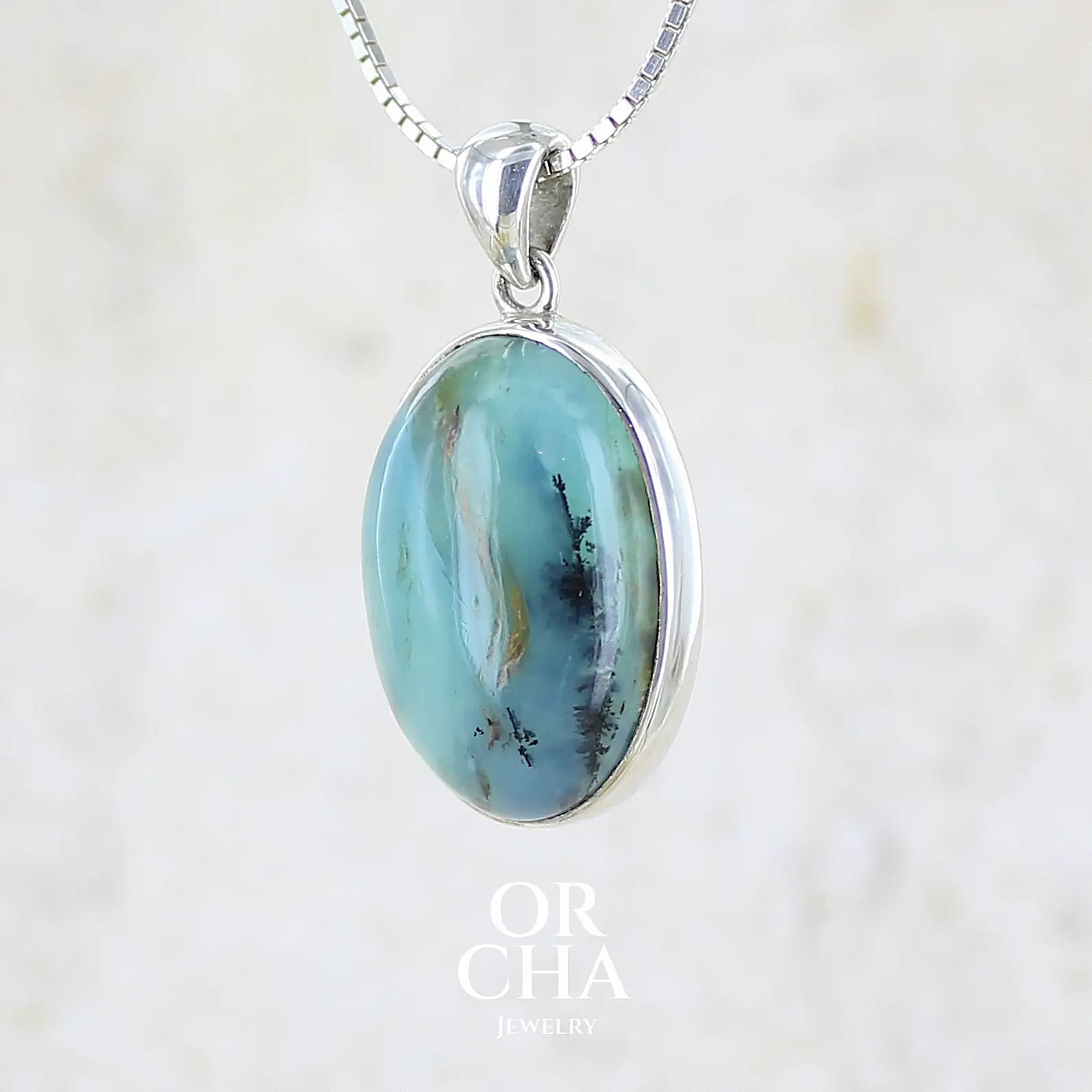 profil gauche d'un pendentif en argent massif, sertie d'un cabochon ovale d'opale bleue du Pérou de couleur bleu laiteux foncé à inclusions de dendrites paysages noires et de roche mère marron. Pierre opaque et translucide. Bijoux unique, éthique et durable présenté dans un coffret de notre marque, prêt à offrir. Livraison offerte, certificat d'authenticité, service client.