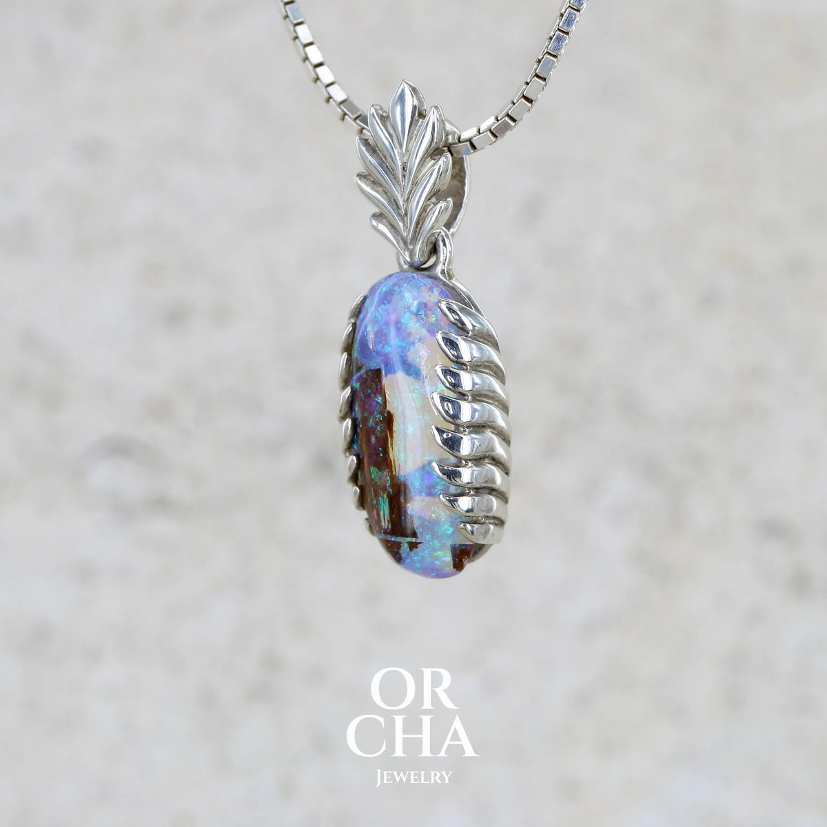Profil gauche d'un pendentif pour femme en argent massif 925 serti d'une opale cristal aux feux étoilés multicolores à dominante bleu violet et vert. Cabochon de 3,2 carats taillé de forme oval de qualité transparente. Bijou unique réalisé par Orchajewelry