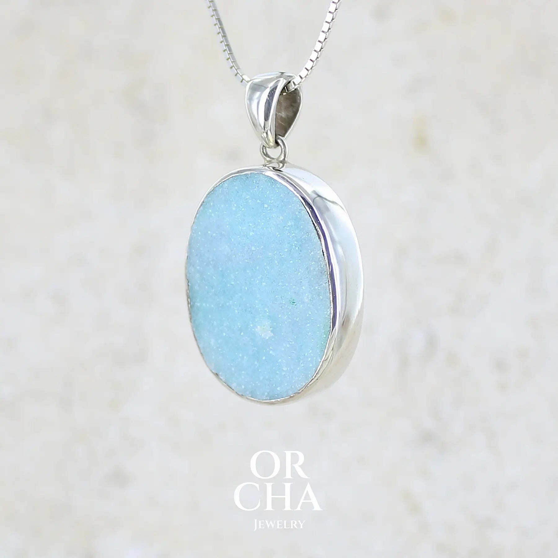 profil gauche d'un pendentif pour femme en argent massif sertie d'un magnifique gemsilica druzy, chrysocolle silicifiée recouverte de fins cristaux de quartz scintillant. Pierre de qualité opaque taillée en tranche de forme ovale. Pierre naturelle originaire de République dominicaine du Congo de couleur bleu clair turquoise. pierre rare et unique
