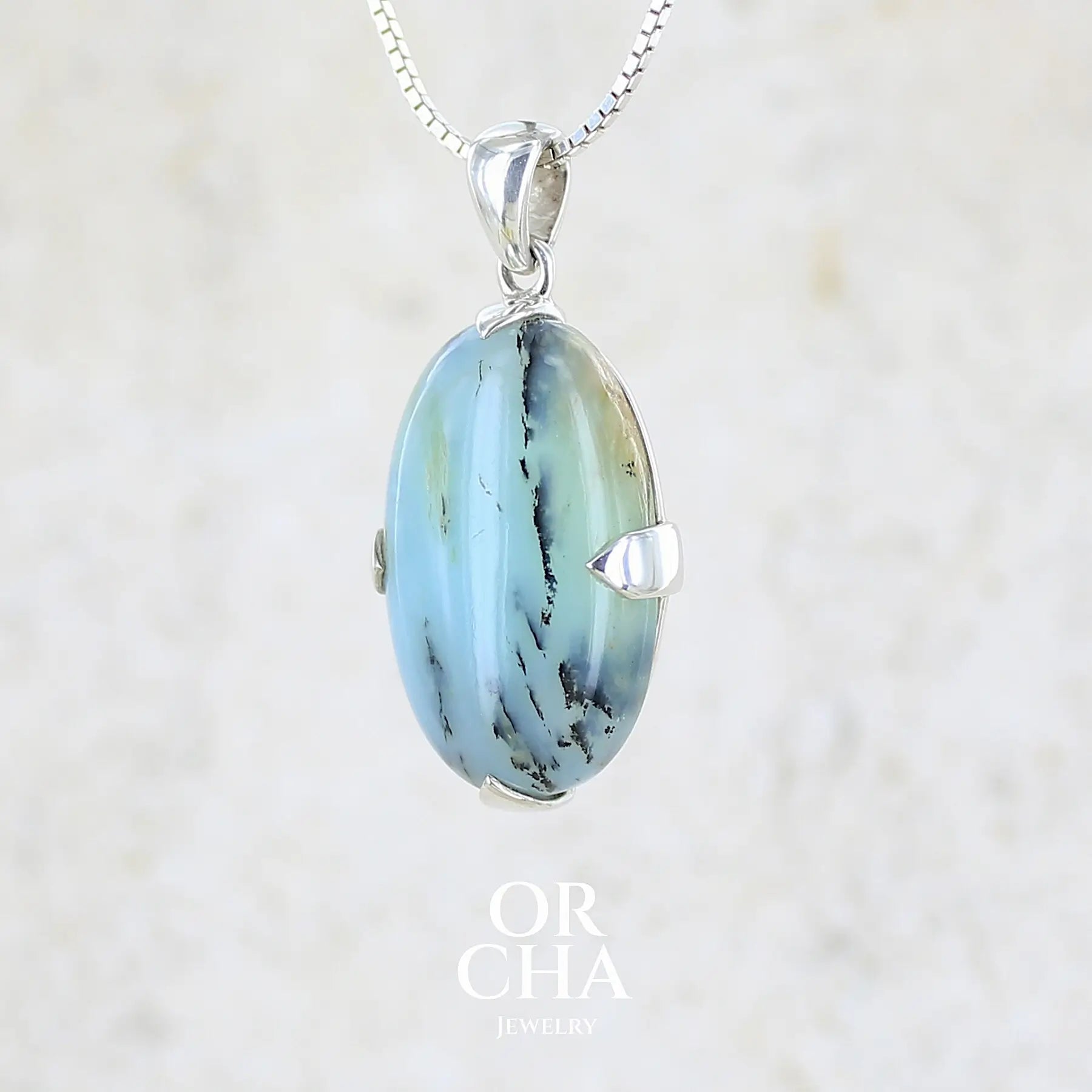 profil gauche d'un pendentif en argent massif, sertie d'un cabochon ovale d'opale bleue du Pérou de couleur bleu laiteux à inclusions de dendrites noires. Pierre opaque et translucide. Bijoux unique, éthique et durable présenté dans un coffret de notre marque, prêt à offrir. Livraison offerte, certificat d'authenticité, service client.