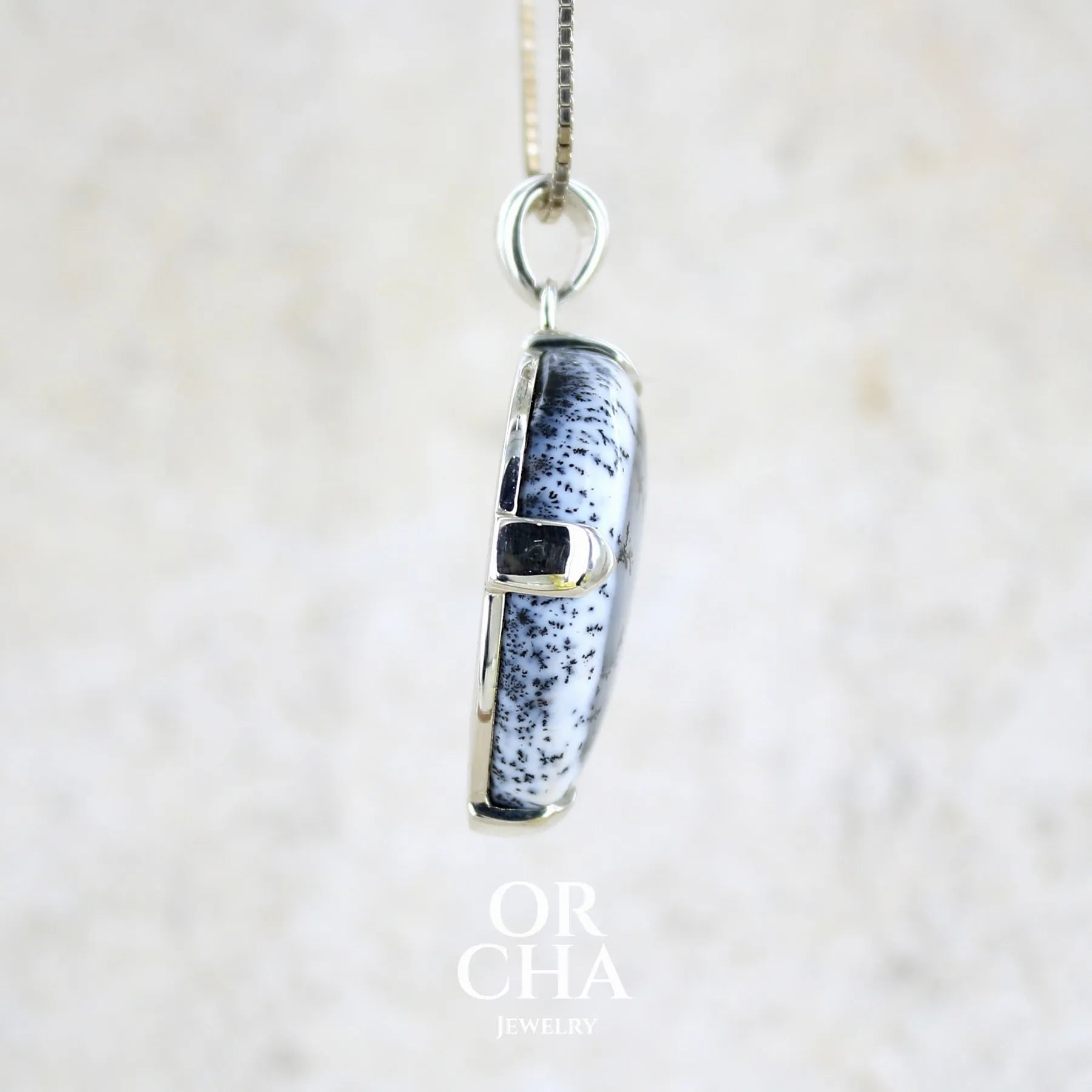 Profil d'un pendentif pour femme en argent massif sertie d'un cabochon en forme de goutte inversée d'agate dendritique de couleur blanc pur et noir. Pierre opaque à translucide à inclusions paysages de dendrites de manganèse noires contrastées. Bijou unique réalisée par Orchajewelry