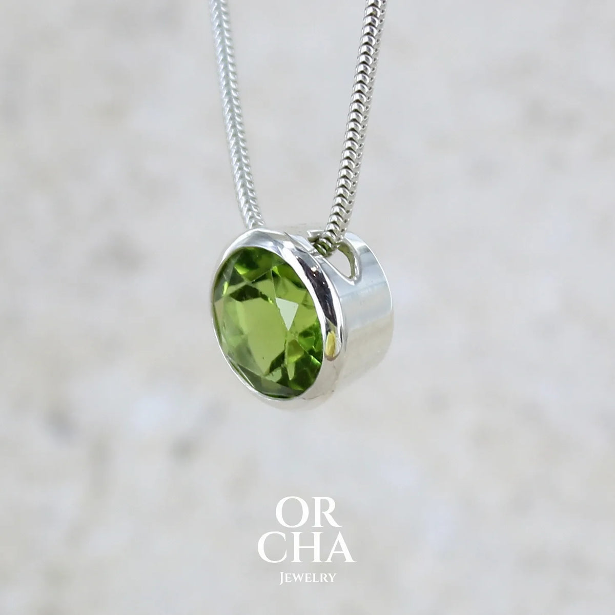 profil gauche d'un collier solitaire pour femme en argent massif serti d'un Péridot facettée de 4 carats. Pierre de forme ronde de qualité transparente, propre à l'oeil. Pierre naturelle de couleur vert olive doré intense et lumineuse. Pendentif monté sur une chaine en argent sterling de type maille serpent de 0,43 mm d'épaisseur et de 40 cm de longueur. Bijoux unique réalisé par Orchajewelry.