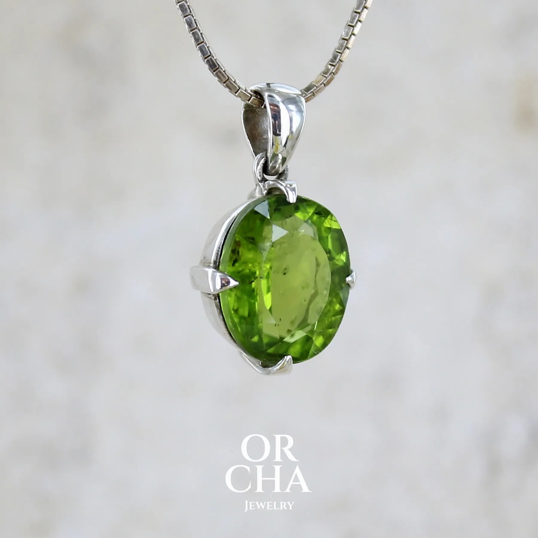 Profil d'un pendentif pour femme en argent massif serti d'un péridot de couleur vert olive doré intense. Pierre taillée, de forme ovale, transparente présentant de nombreuse inclusions. Bijou unique réalisé par Orchajewelry
