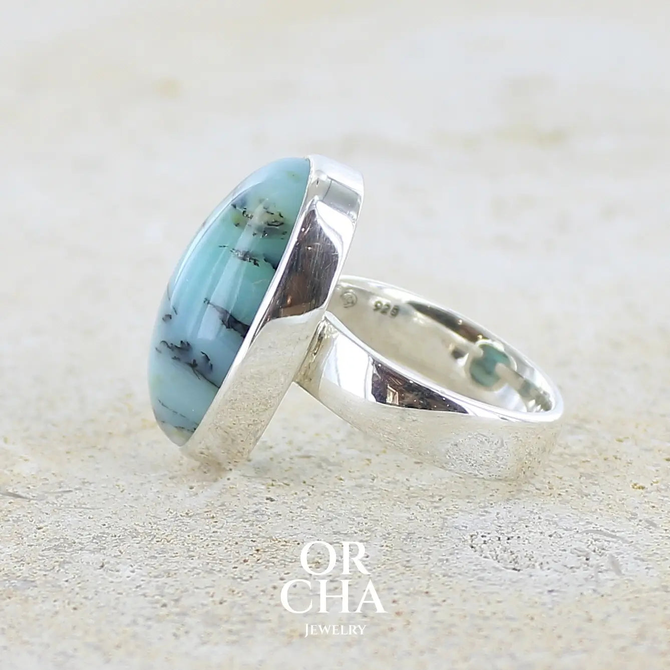 Détail sertissage d'une bague pour femme en argent massif, sertie d'un cabochon ovale d'opale bleue du Pérou de couleur bleu laiteux à inclusions de dendrites noires. Pierre naturelle de qualité opaque et translucide. Bijoux unique réalisé en taille 54,5 - OrchaJewelry