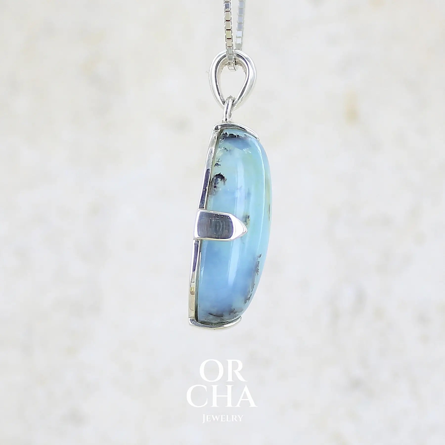 détail de texture d'un pendentif pour femme et homme en argent massif, sertie d'un cabochon d'opale bleue du Pérou de couleur verte bleutée. Pierre en forme de goutte inversée. Pierre opaque et translucide. Bijoux unique, éthique et durable présenté dans un coffret de notre marque, prêt à offrir. Livraison offerte, certificat d'authenticité, service client.