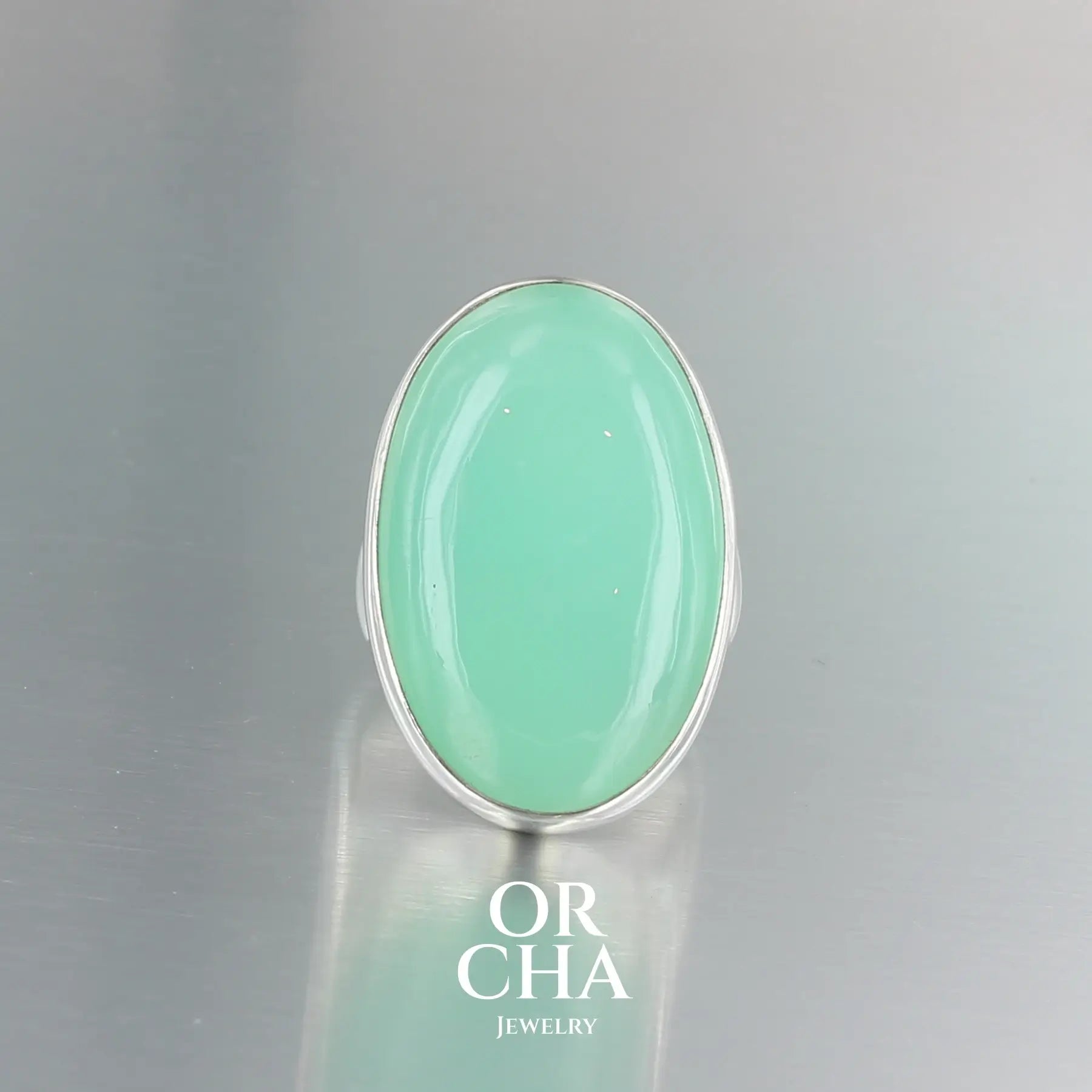 Grande bague contemporaine pour femme en argent massif sertie d'une Chrysoprase. Pierre cabochon de forme ovale de qualité opaque à translucide. Pierre naturelle de couleur vert bleuté lumineux. réalisée en taille 58. Bijoux unique, éthique et durable présenté dans un coffret de notre marque, prêt à offrir. Livraison offerte, certificat d'authenticité, service client.